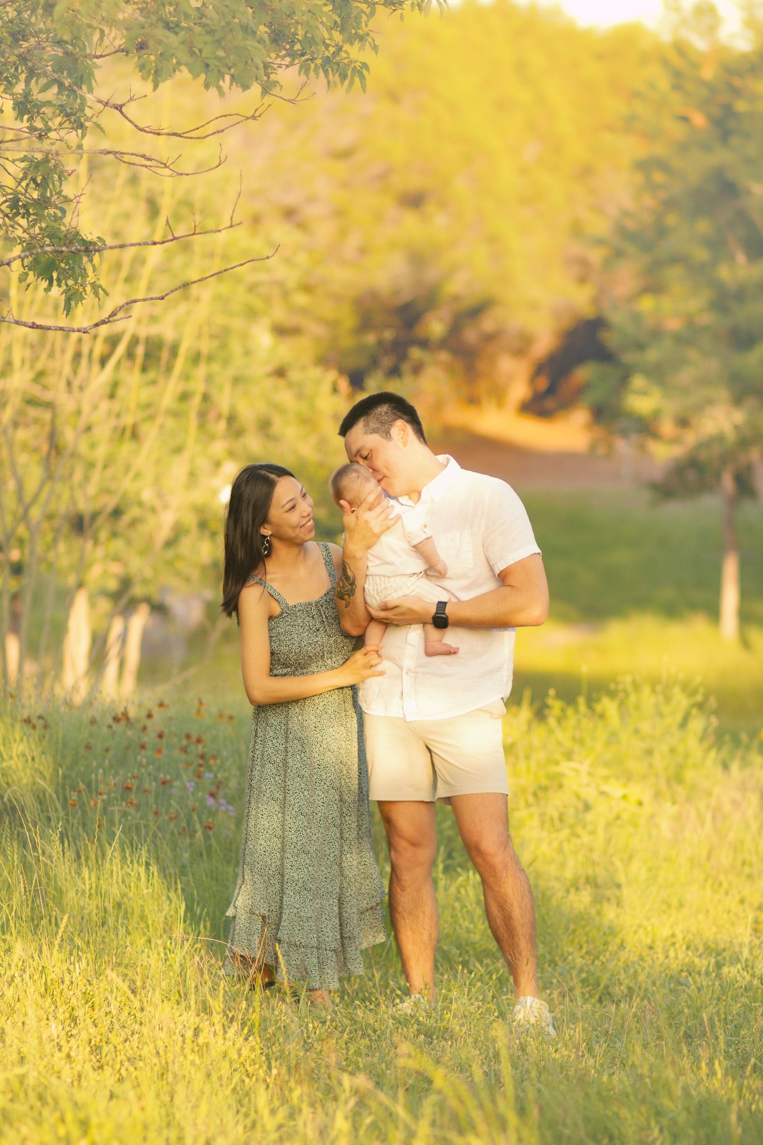 Young family with baby sharing tender moment in lush green field at sunset.