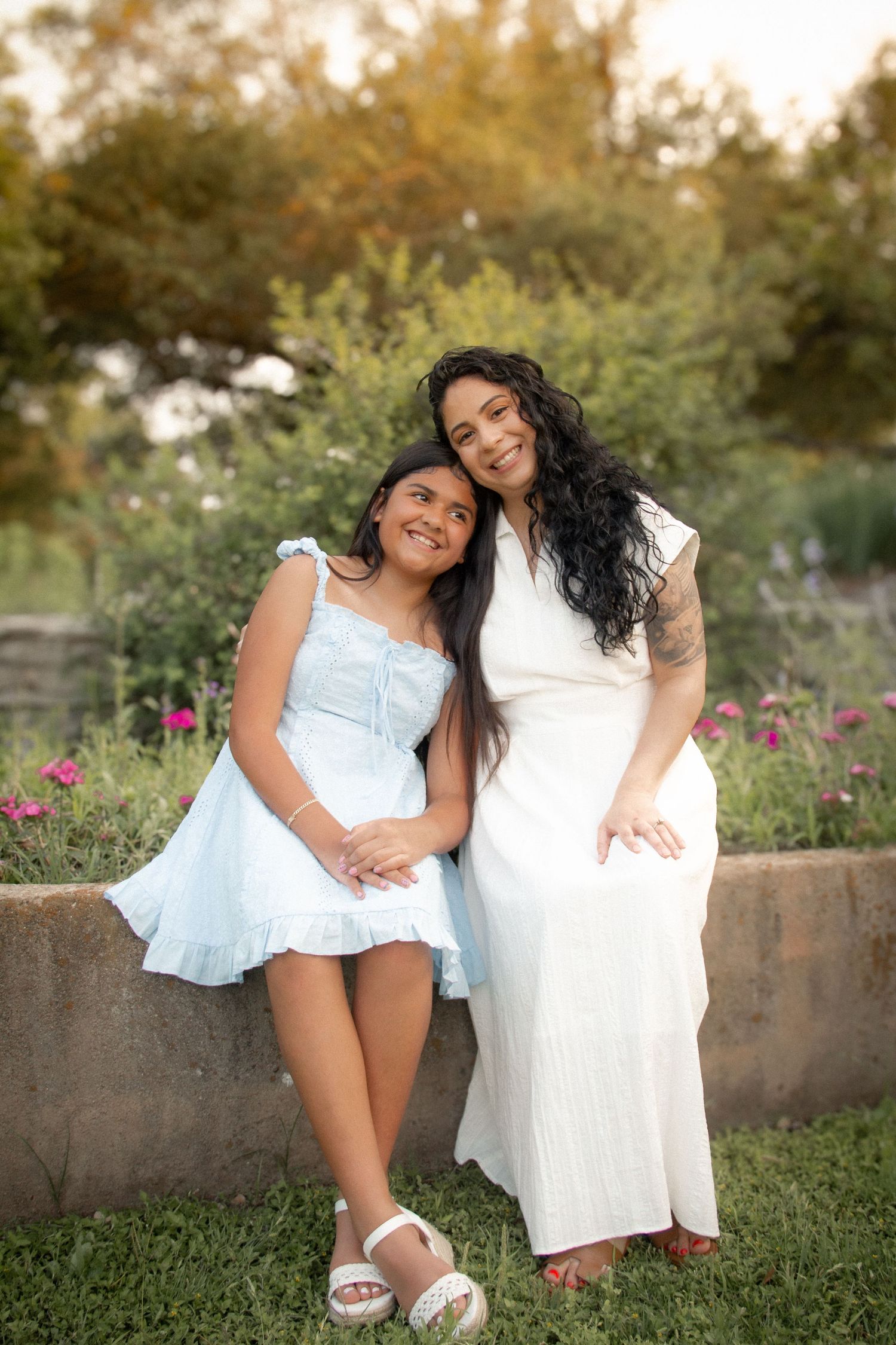 A woman and young girl in white and blue dresses smile together in a lush garden setting.