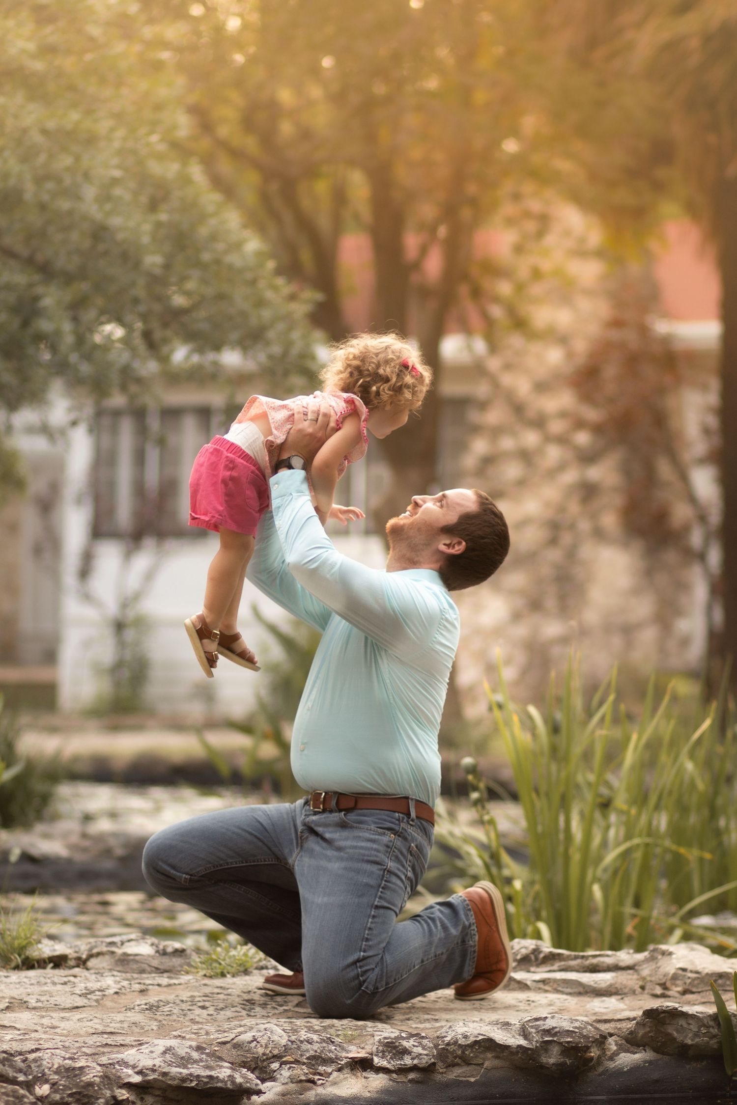 Father kneeling outdoors, joyfully lifting his curly-haired toddler daughter in a pink dress above his head.