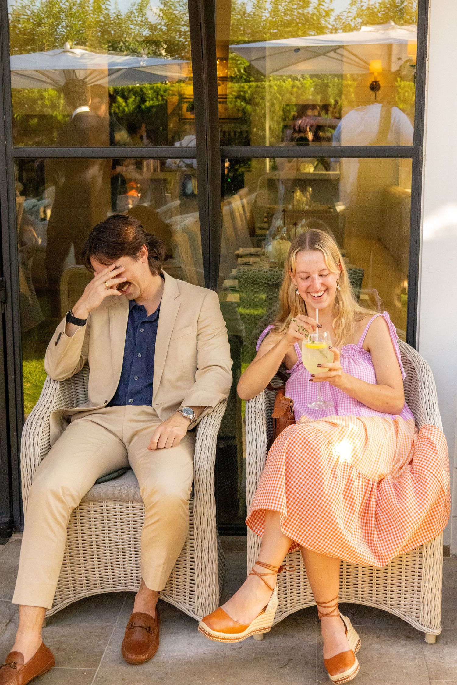 Man in beige suit and woman in pink dress laughing while sitting in wicker chairs.