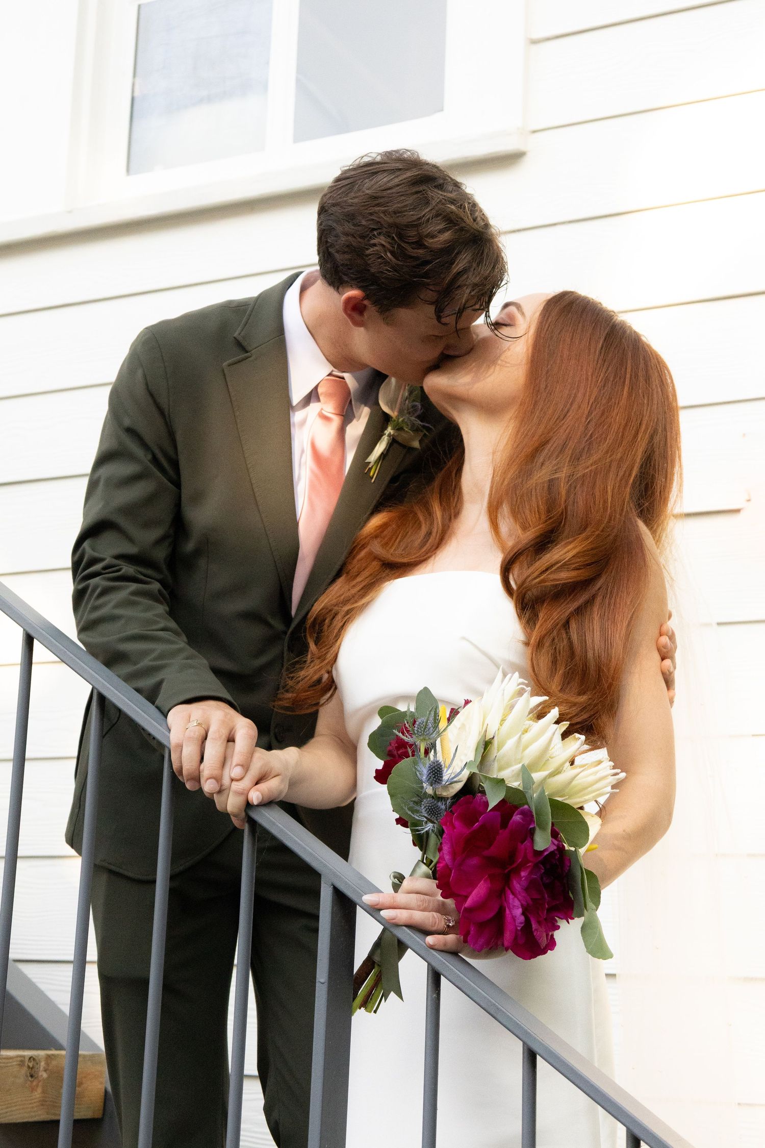 Bride and groom kissing on stairs, bride holding burgundy bouquet, groom in olive suit with coral tie.