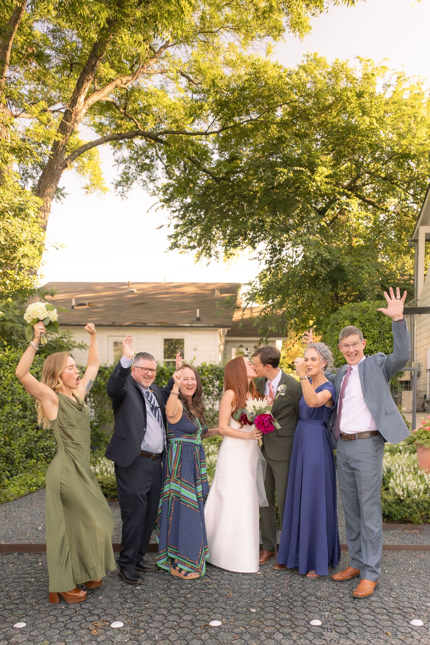 A joyful wedding party celebrates outdoors as the bride and groom share a kiss, surrounded by cheering family members.