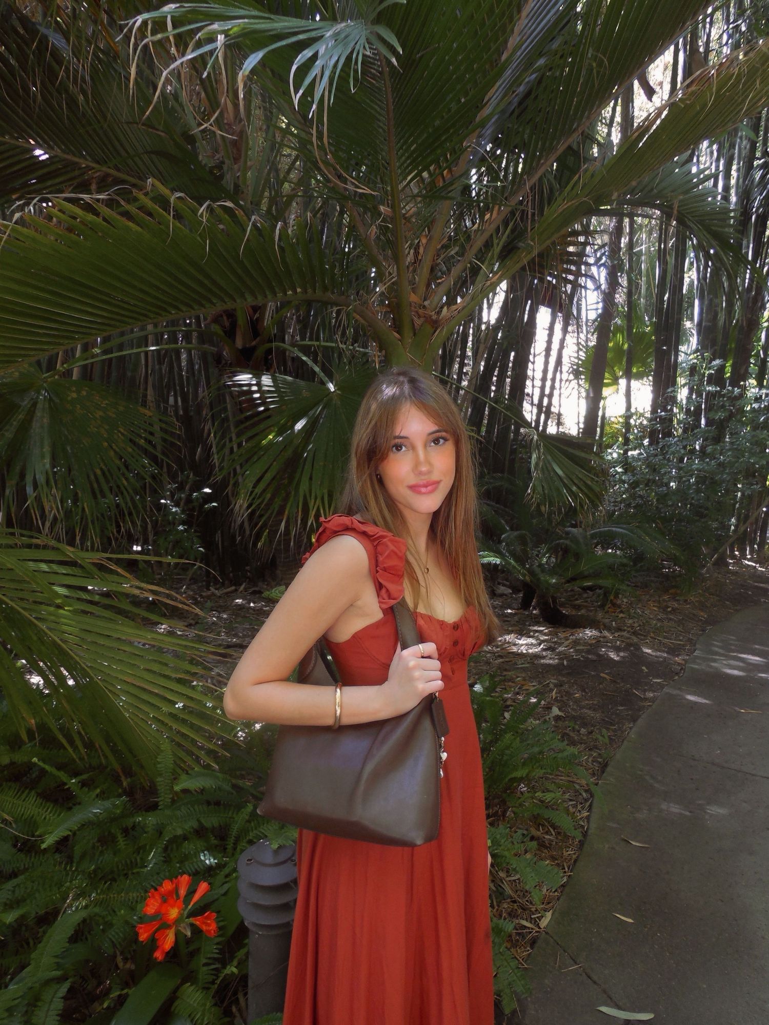 A young woman in a red dress carries a dark handbag while standing among lush tropical palm trees.