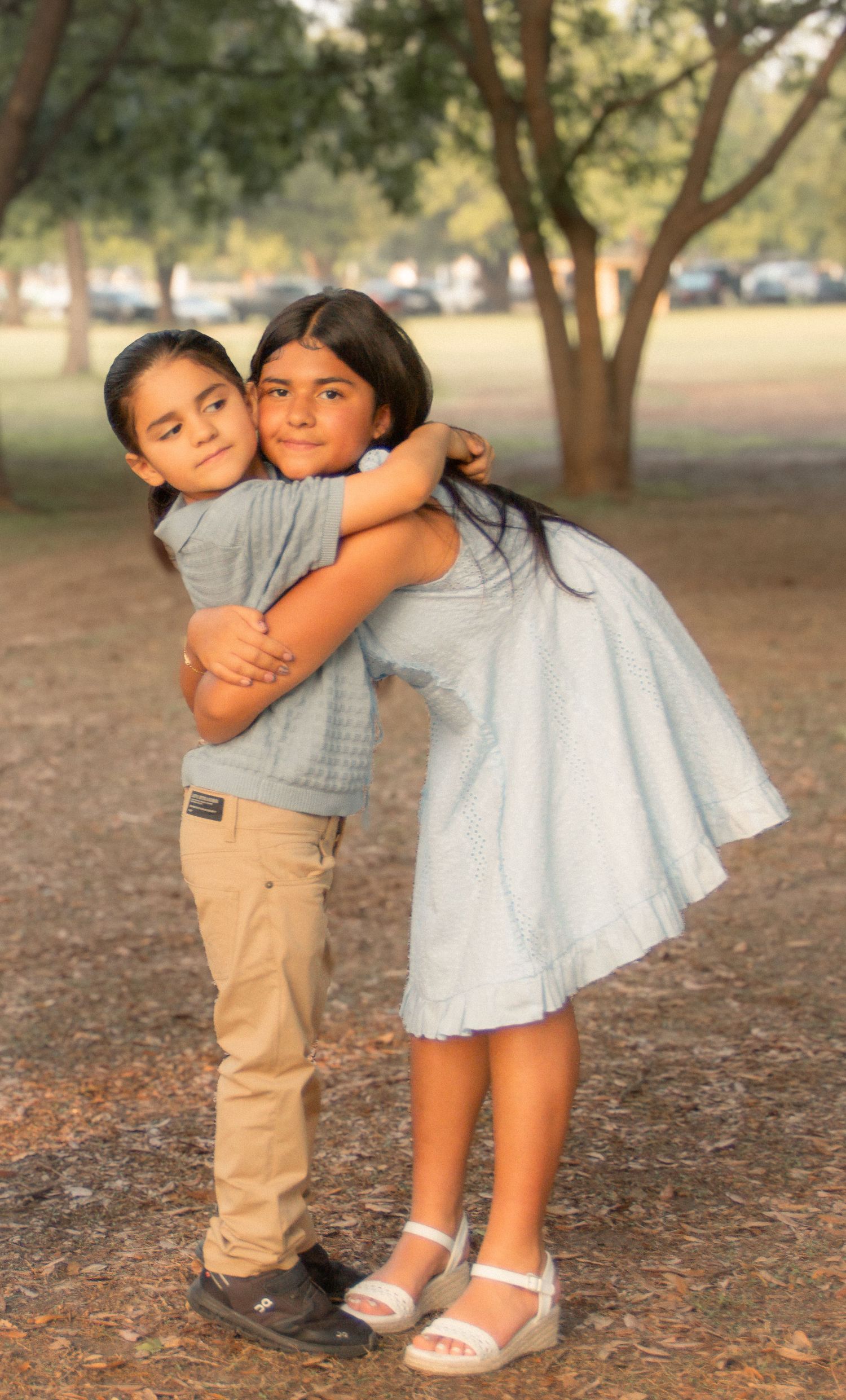Two children hugging warmly outdoors in a park, a boy in khakis and a girl in a light blue dress, surrounded by trees.