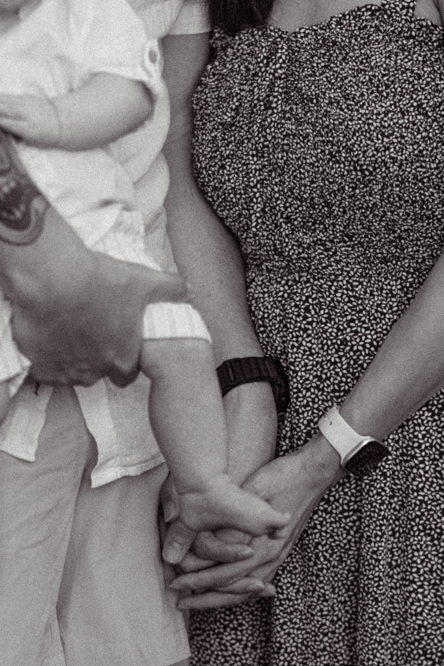 Black and white close-up of couple holding hands while father carries newborn baby, woman in polka dot dress.