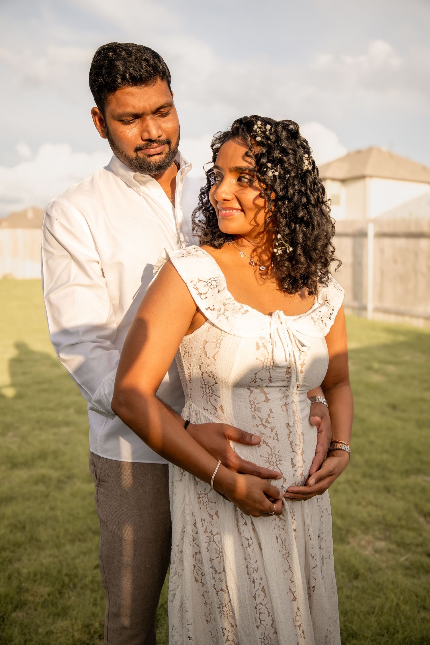 A pregnant woman in a white lace dress stands with her partner outdoors, both cradling her baby bump in golden sunlight.