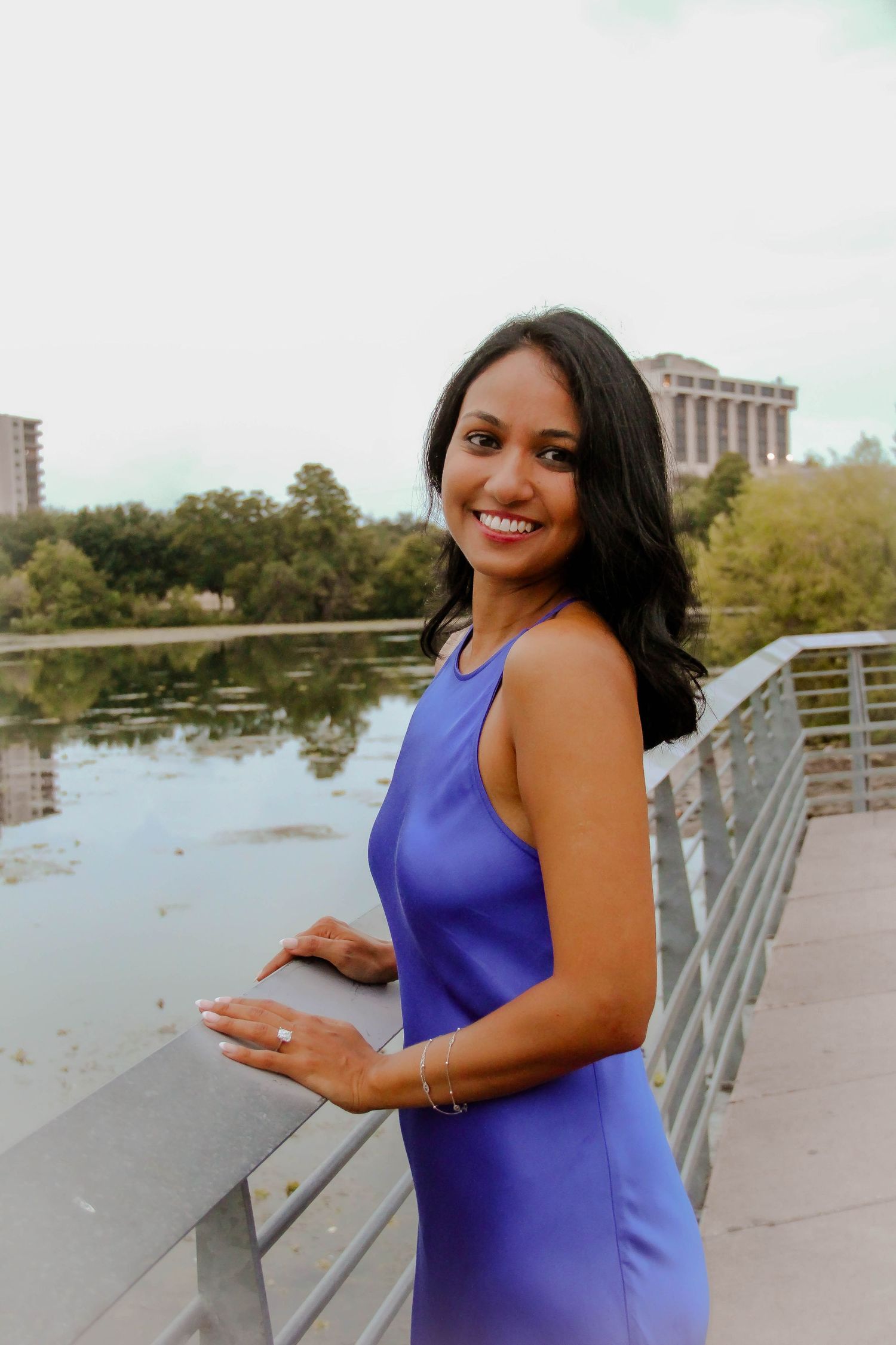 Smiling woman in blue dress displaying engagement ring on a bridge by a lake.