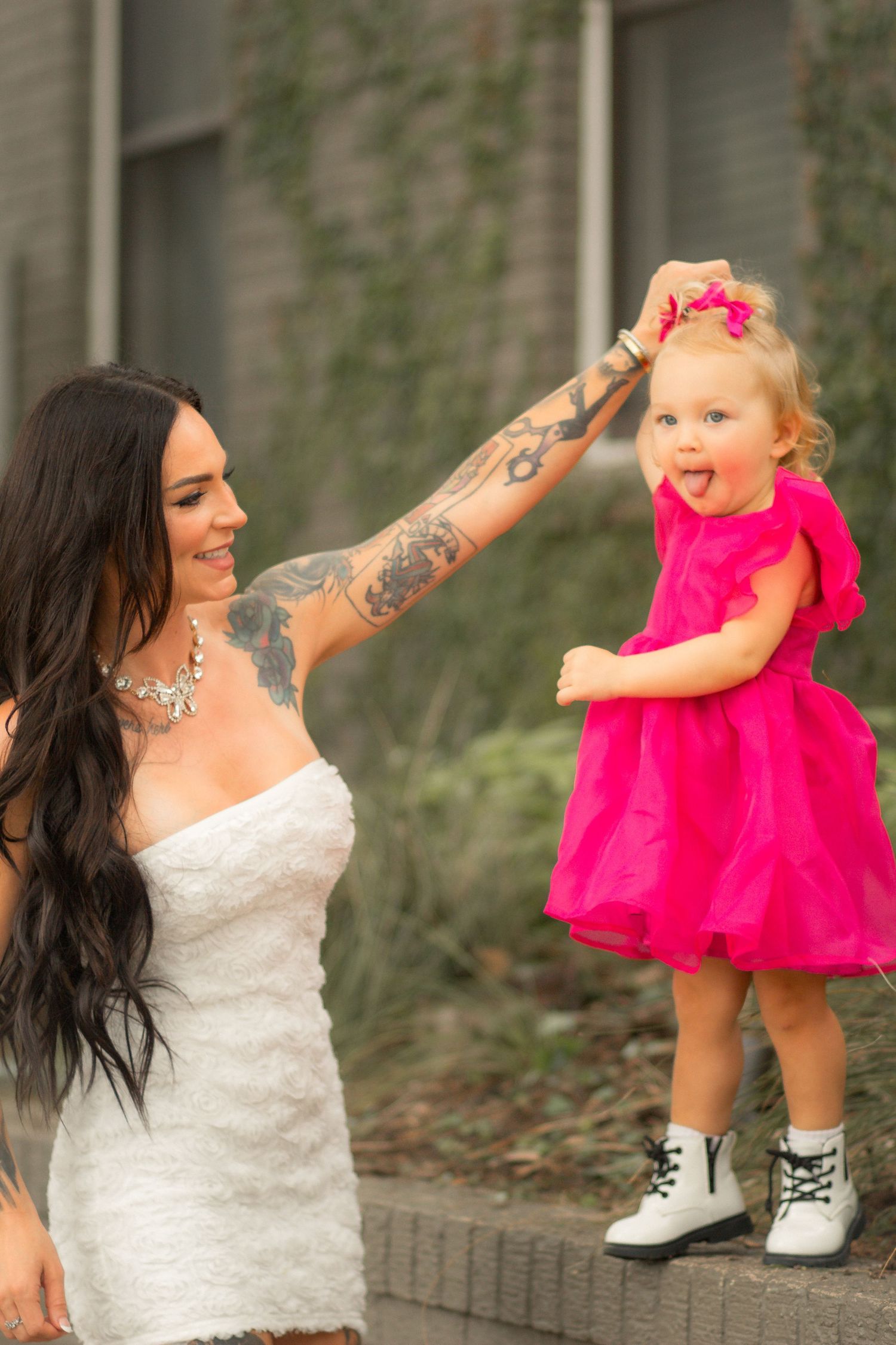 A tattooed woman in a white dress twirls a toddler girl in a pink dress and white boots outdoors.