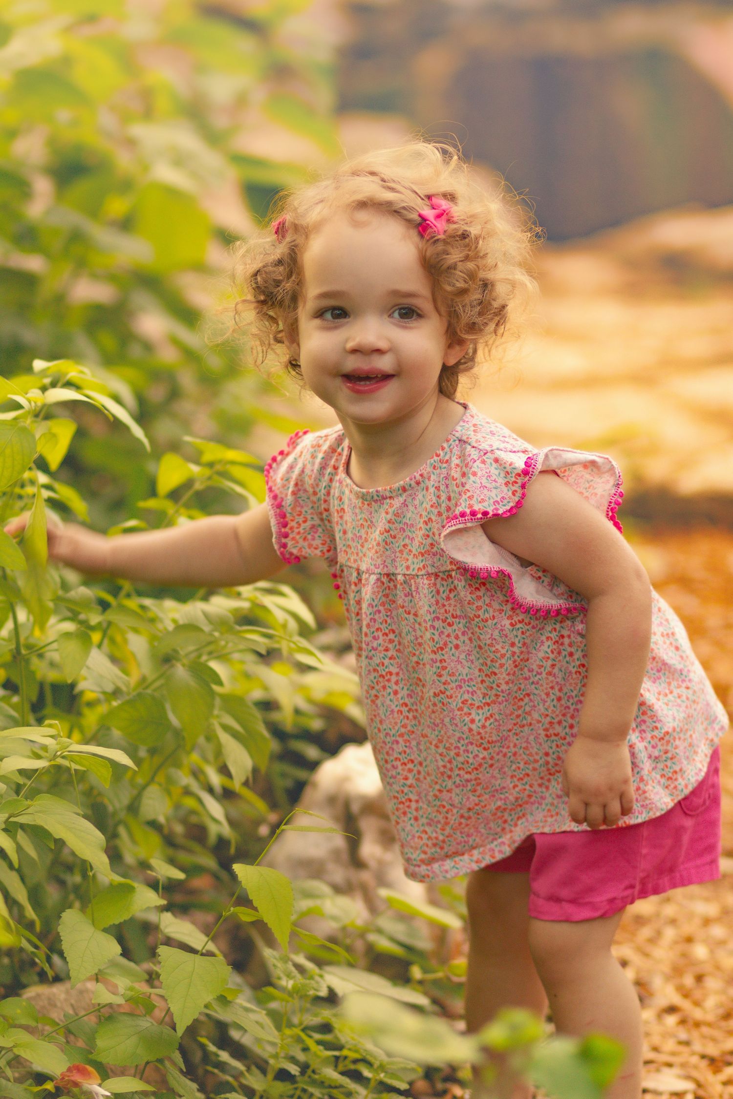 Smiling toddler girl with curly hair in pink outfit touching green plants outdoors.