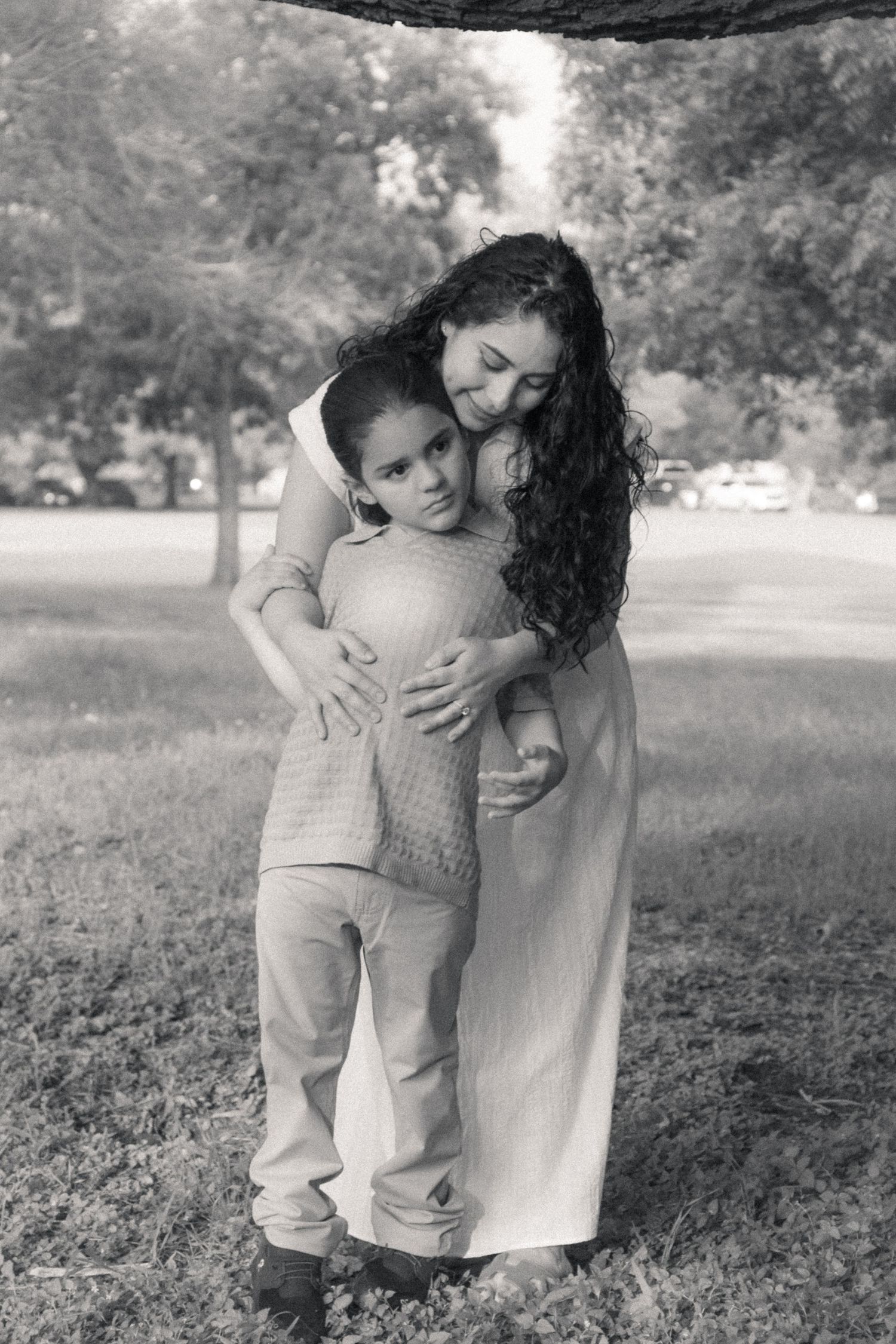 Black and white photo of a mother tenderly hugging her young son in a park setting.