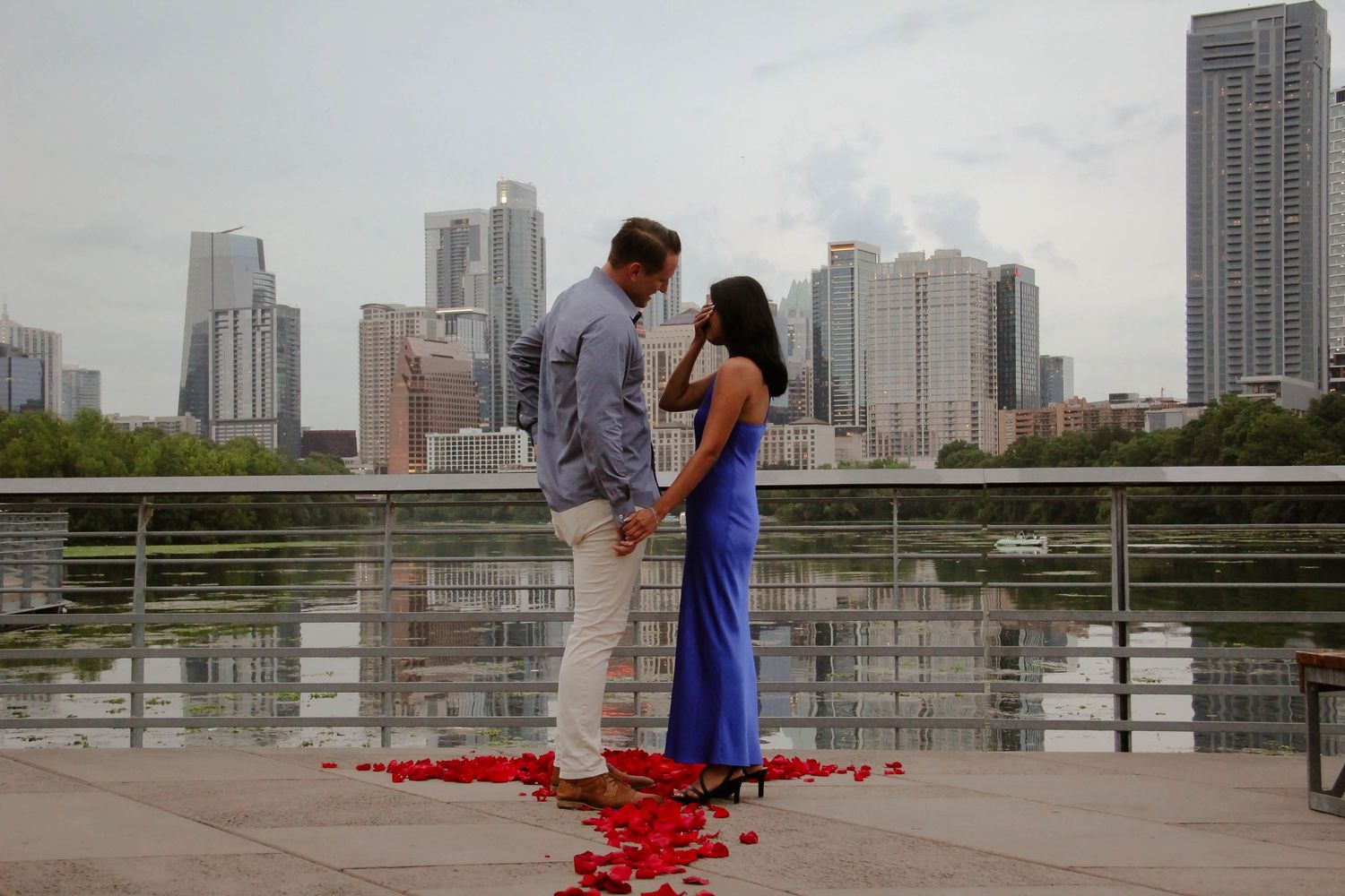Newly engaged couple standing face to face surrounded by red rose petals by city skyline.
