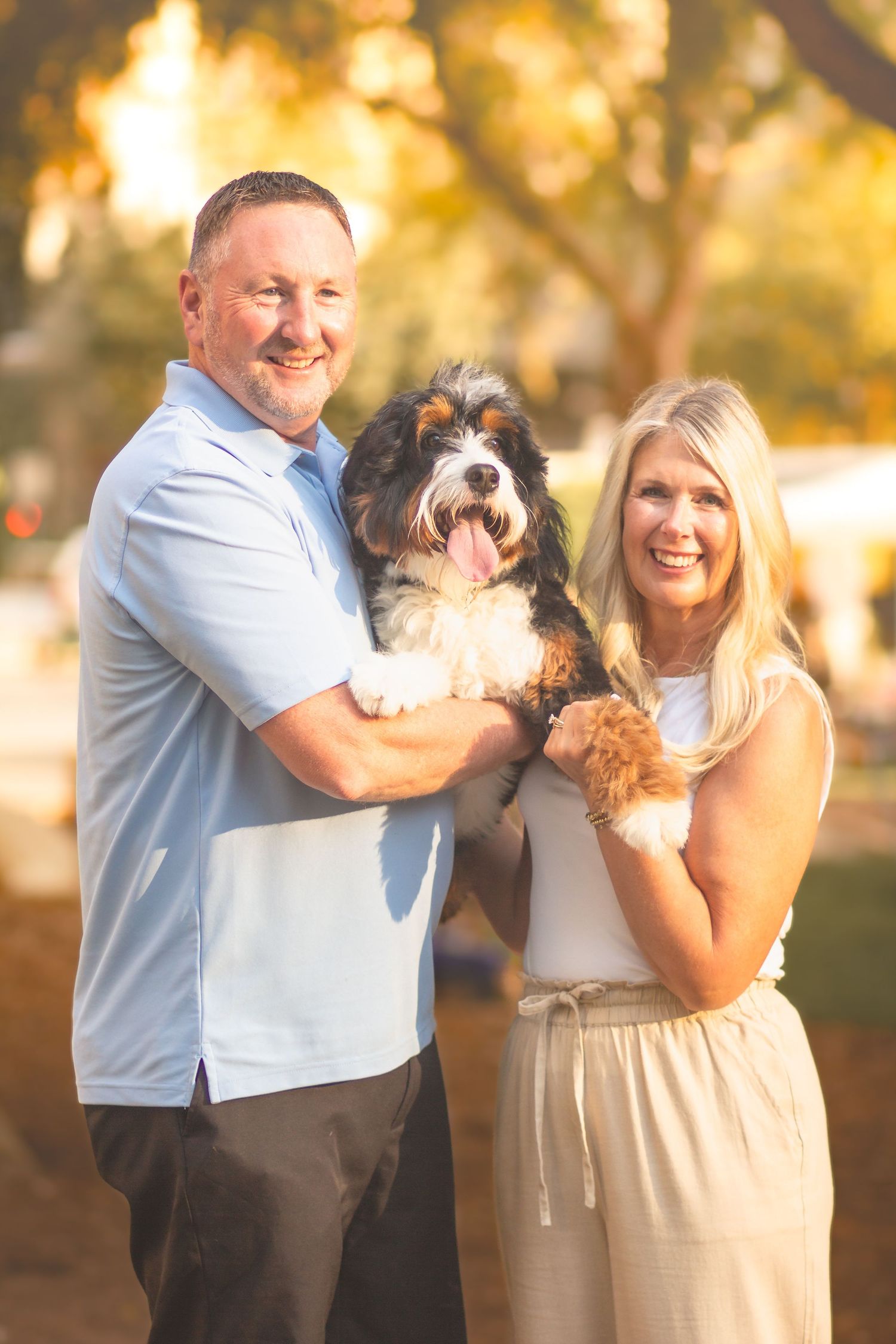 A smiling couple holds a fluffy tricolor dog outdoors in warm golden light, surrounded by blurred trees.