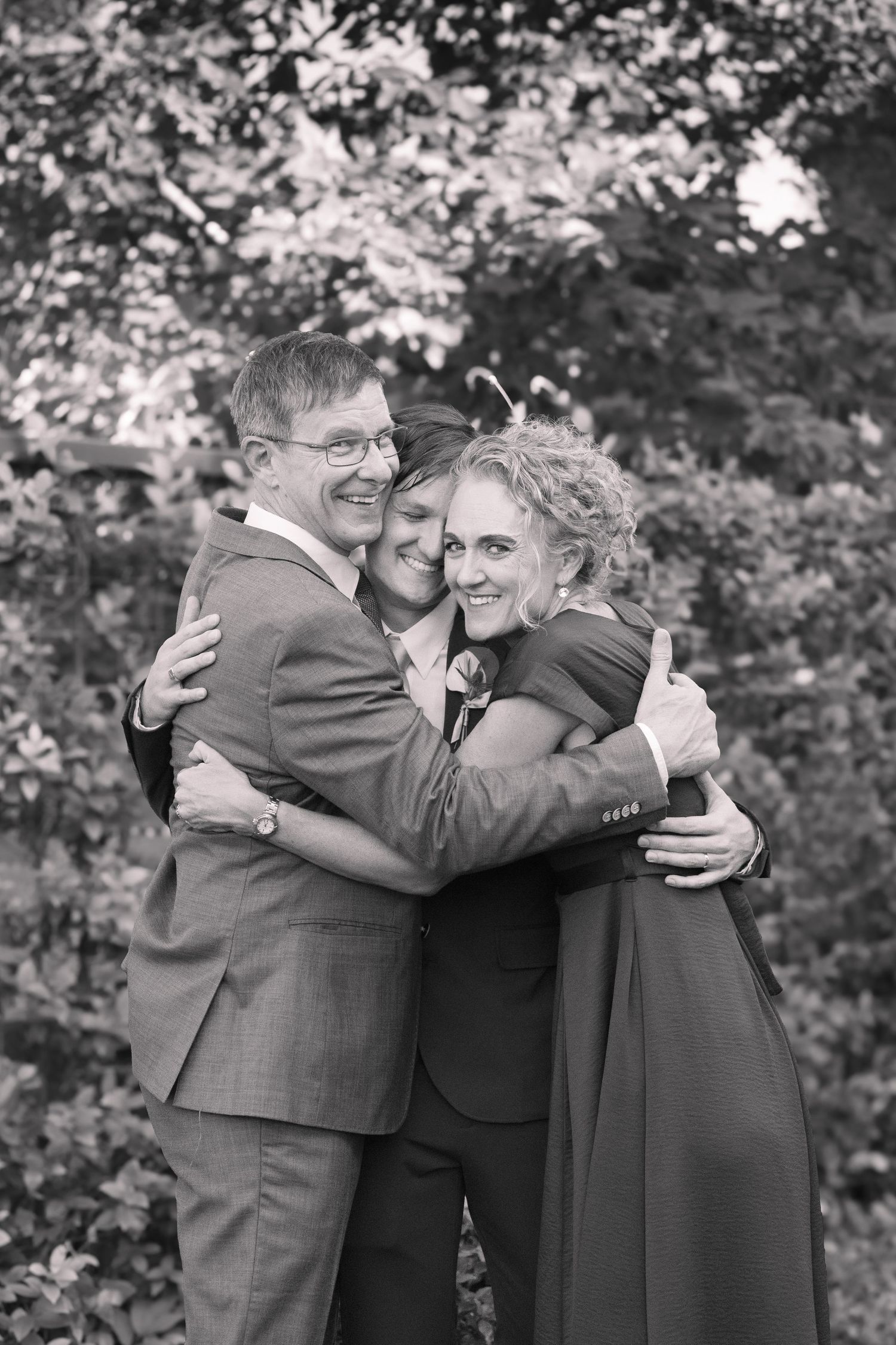 Black and white photo of a bride and groom sharing a warm group hug with a woman outdoors at a wedding.