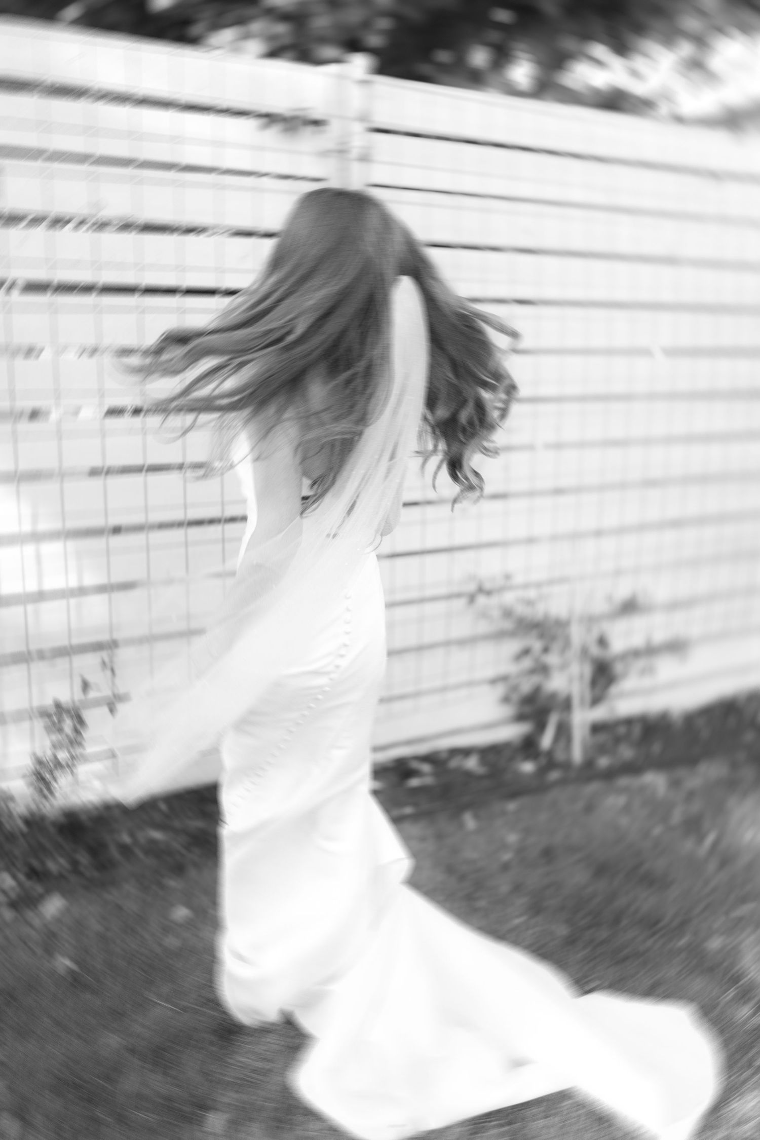 Black and white photo of a bride in a flowing wedding dress spinning near a wooden fence outdoors.