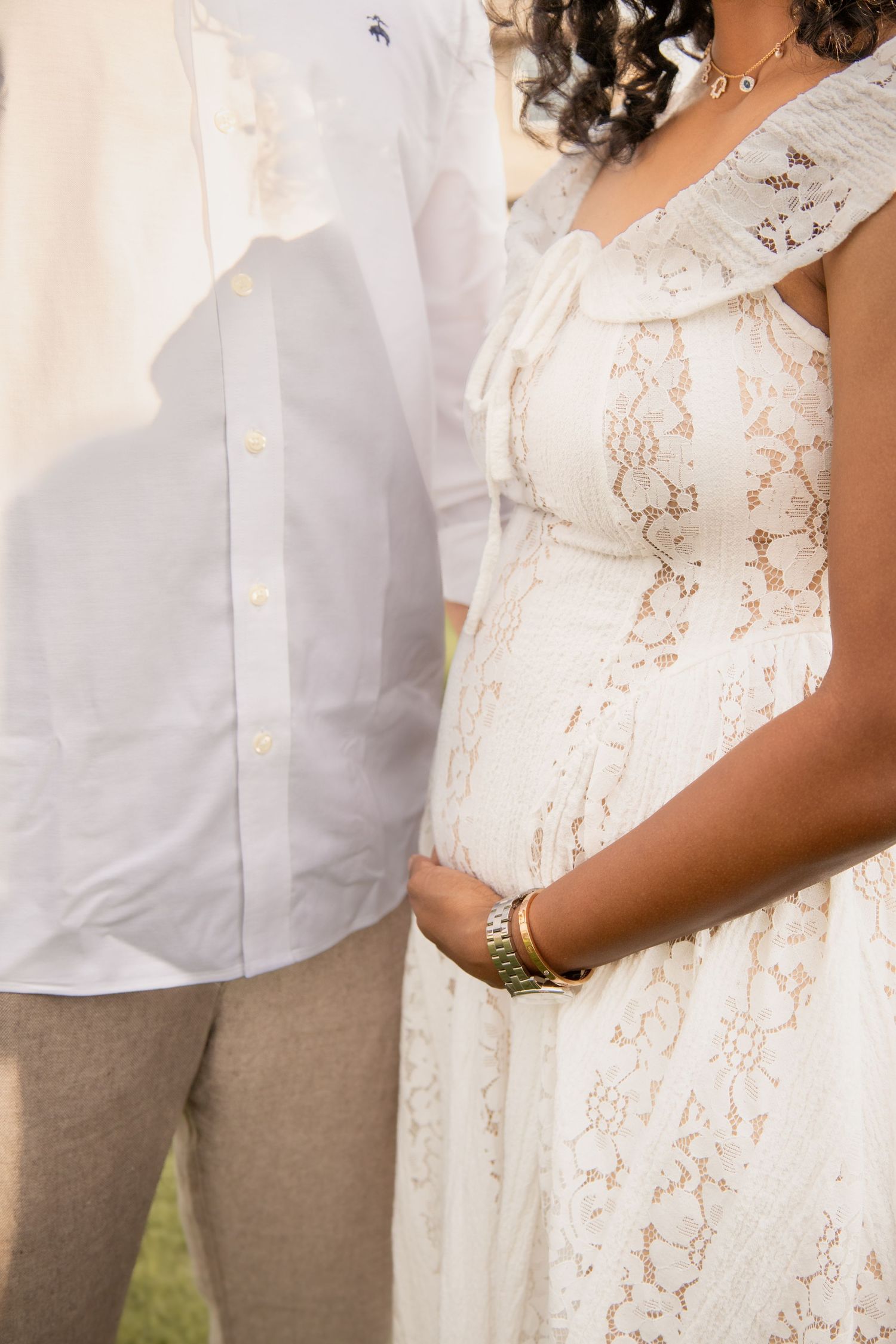 Close-up of a pregnant woman in a white lace dress holding her baby bump, standing beside a man in white shirt.