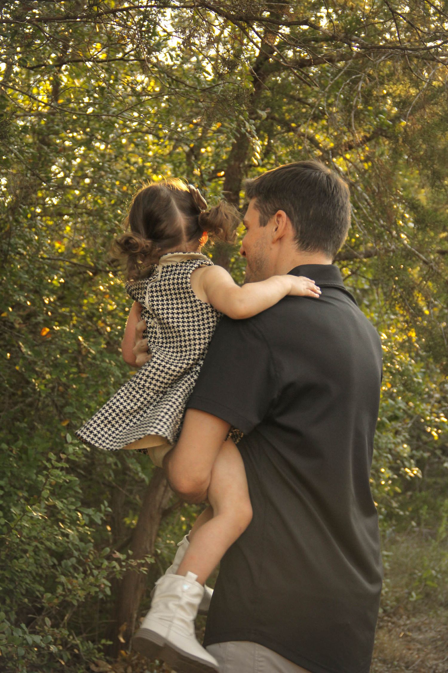 A father holds his young daughter in a checkered dress outdoors among green trees in warm sunlight.