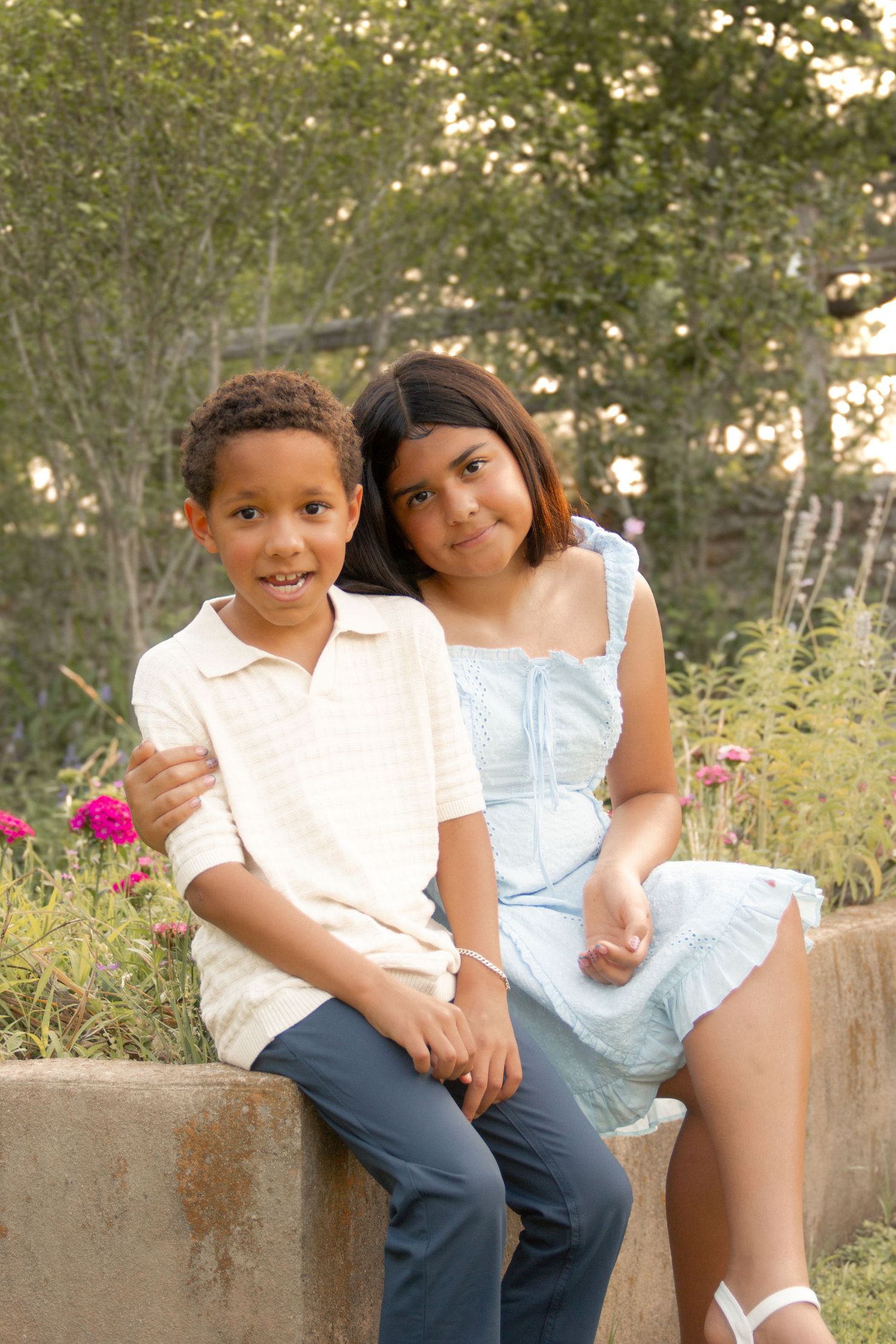 Two smiling children, a boy in white shirt and girl in blue dress, sit together on a stone wall in a garden.