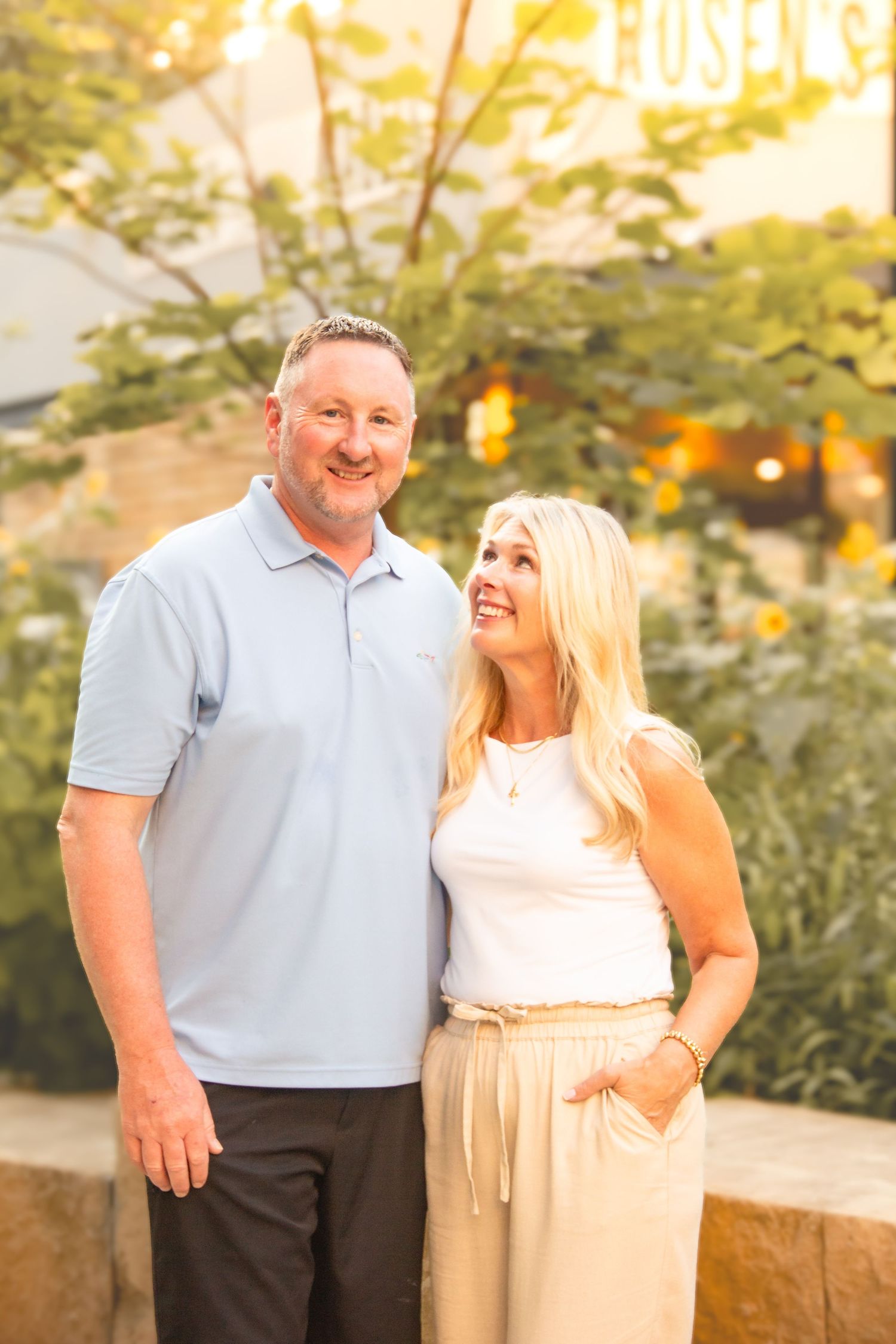 Smiling couple posing outdoors at golden hour, man in light blue polo and woman in white top with beige pants.