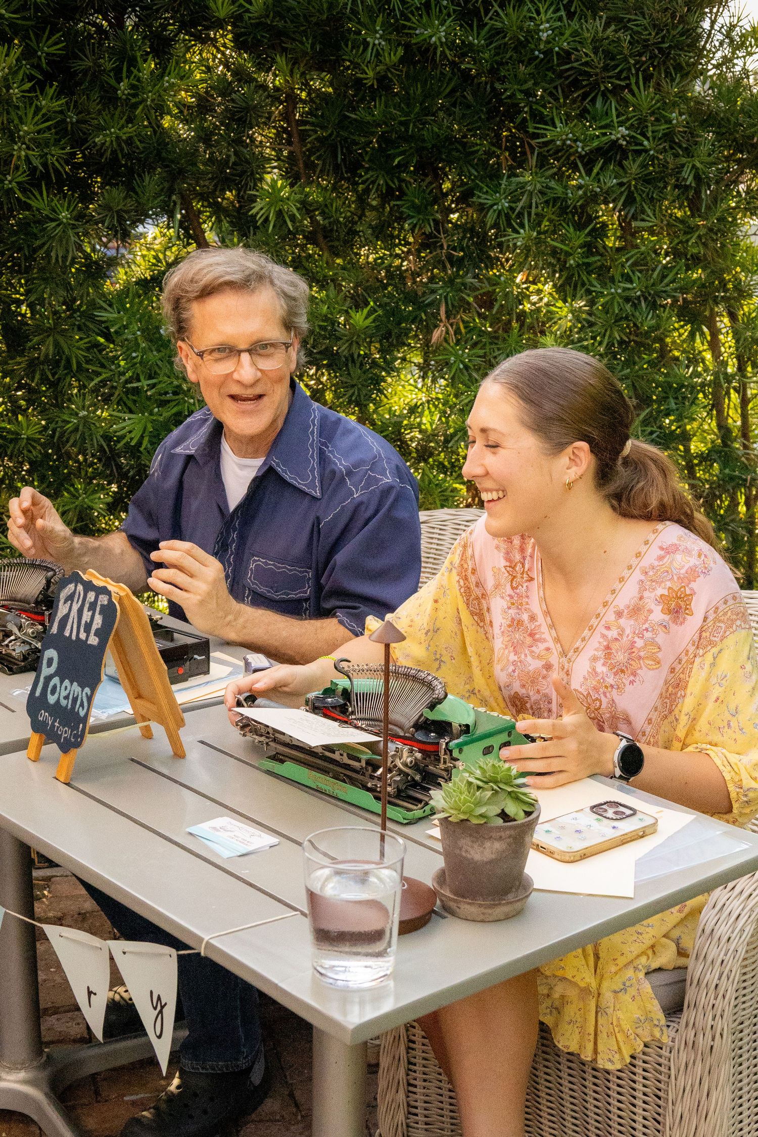 Two people laughing outdoors at a table with a typewriter and a Free Poems sign surrounded by lush greenery.