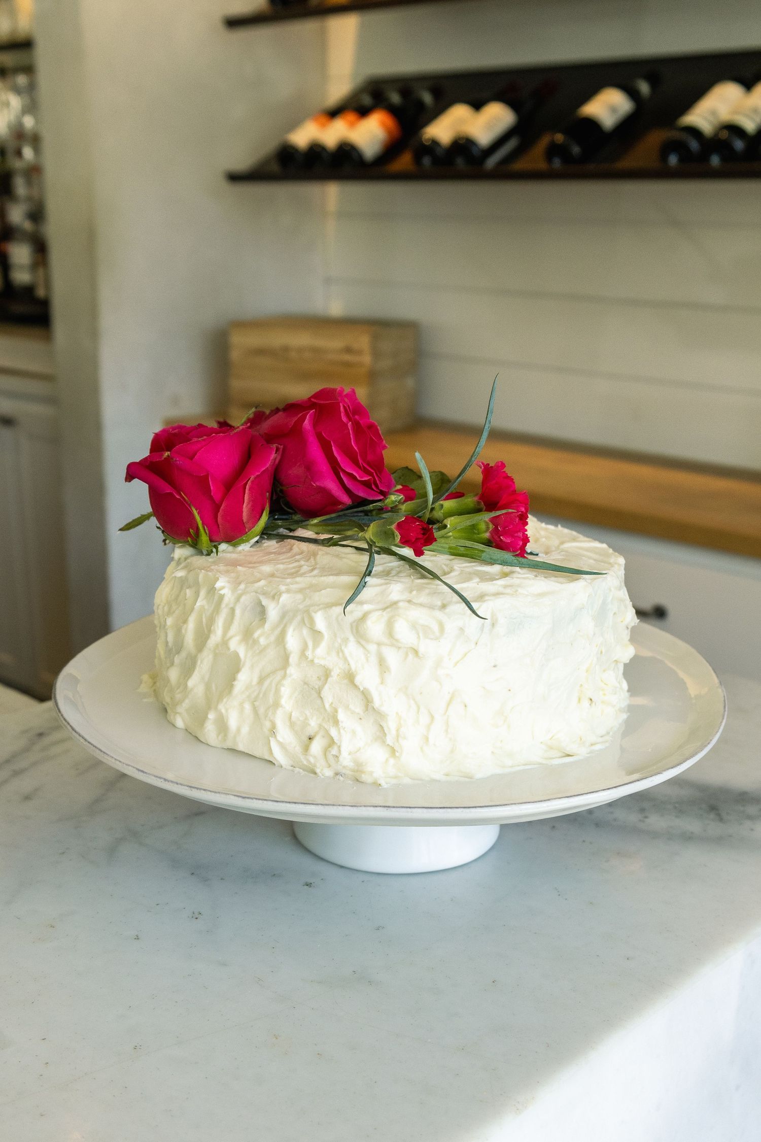White frosted cake topped with red roses on a marble cake stand in an elegant bakery setting.