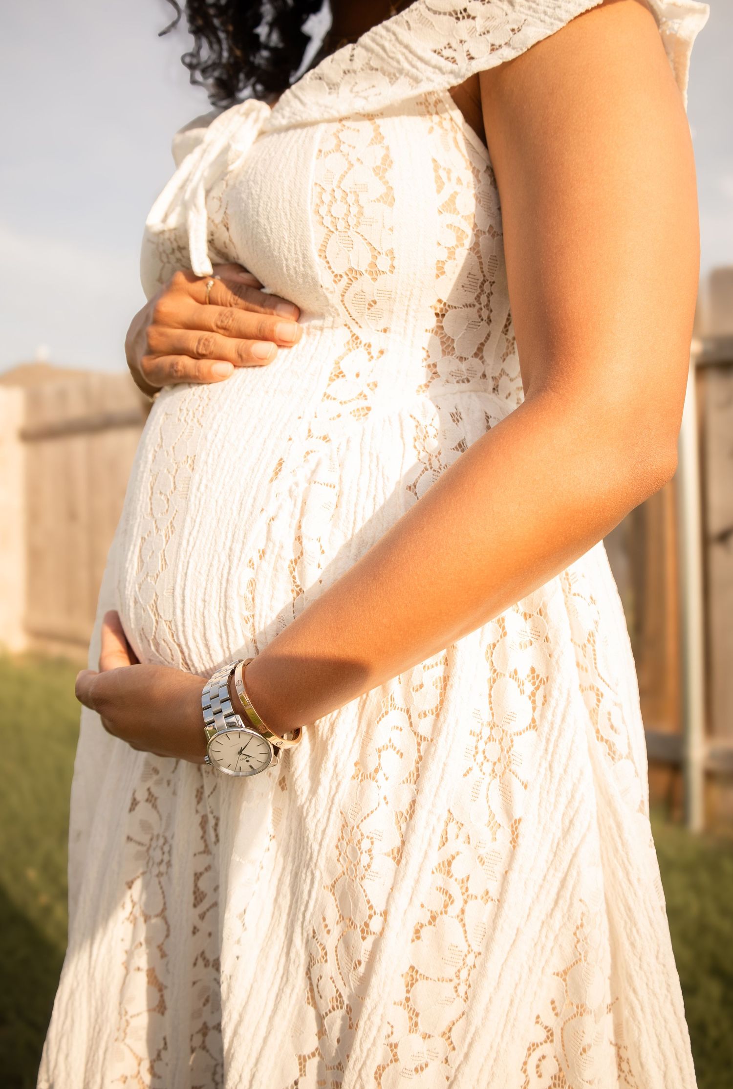 Pregnant woman in white lace dress cradling baby bump outdoors, wearing a silver watch, maternity photography.