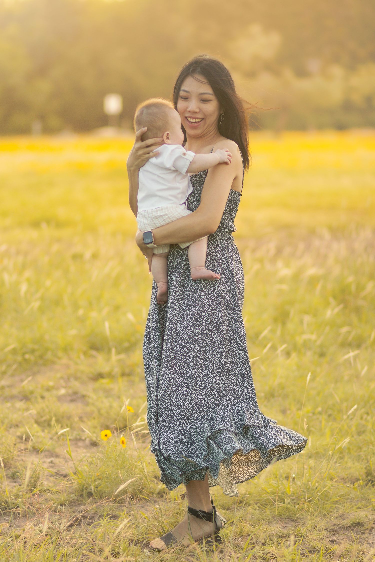 A smiling Asian woman in a floral dress holds her baby in a sunlit field of yellow flowers.