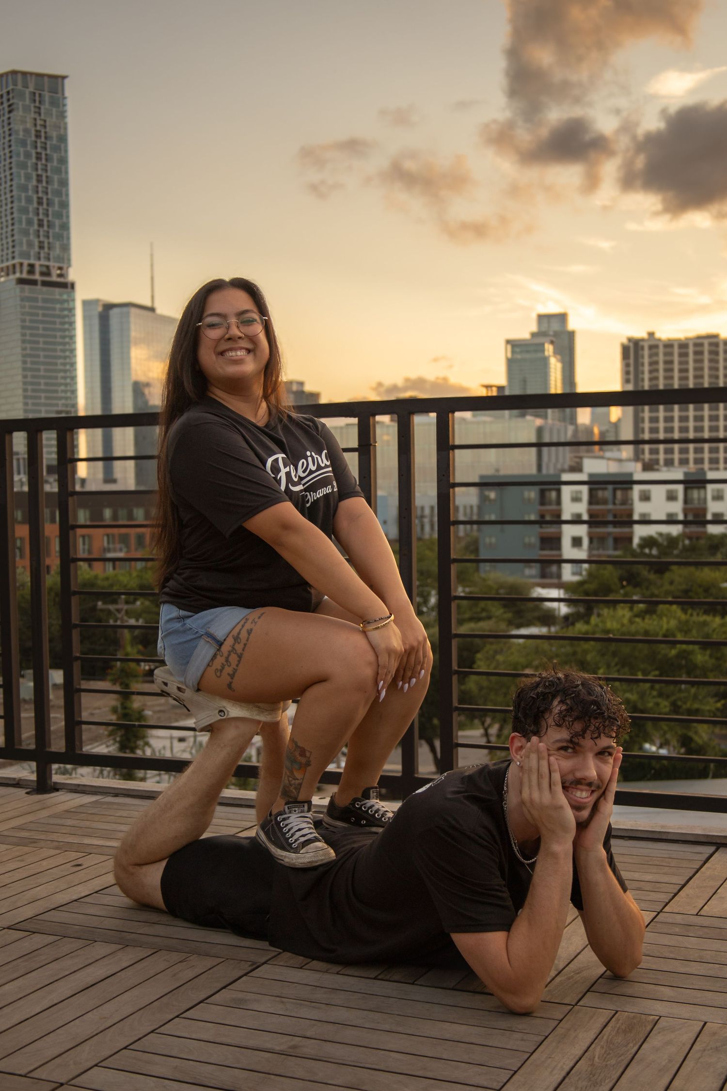 Two friends pose playfully on a rooftop deck at sunset with city skyline views behind them.