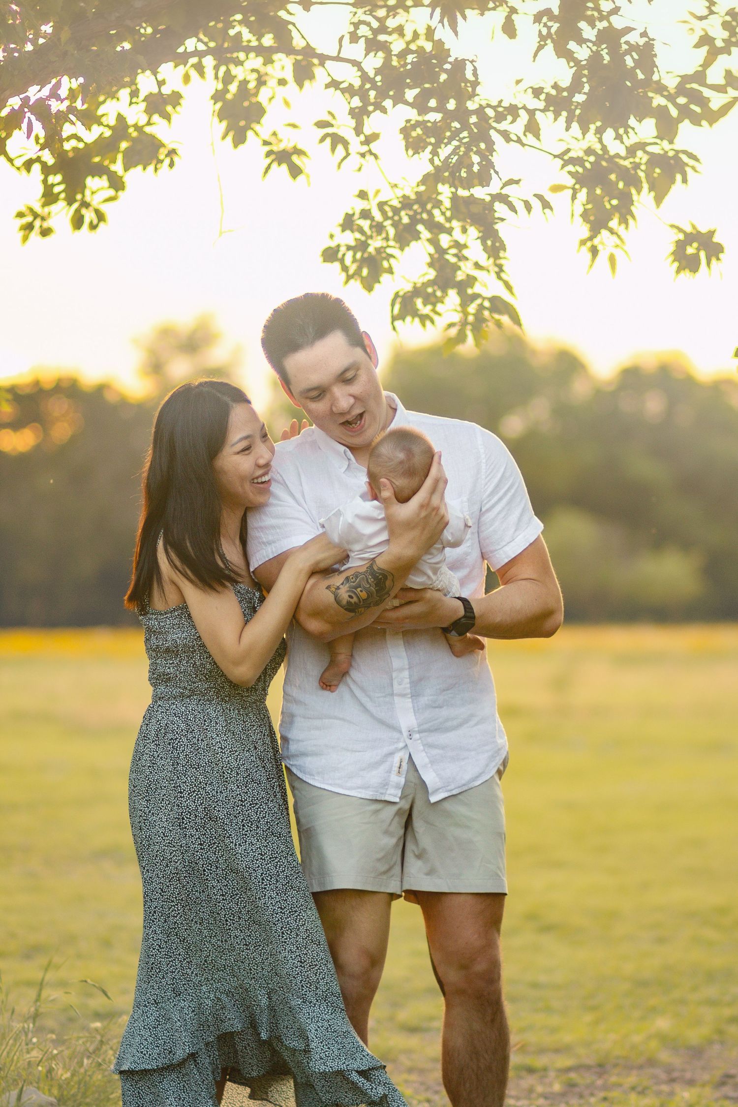 Joyful young family with newborn laughing together under tree in golden sunset field.