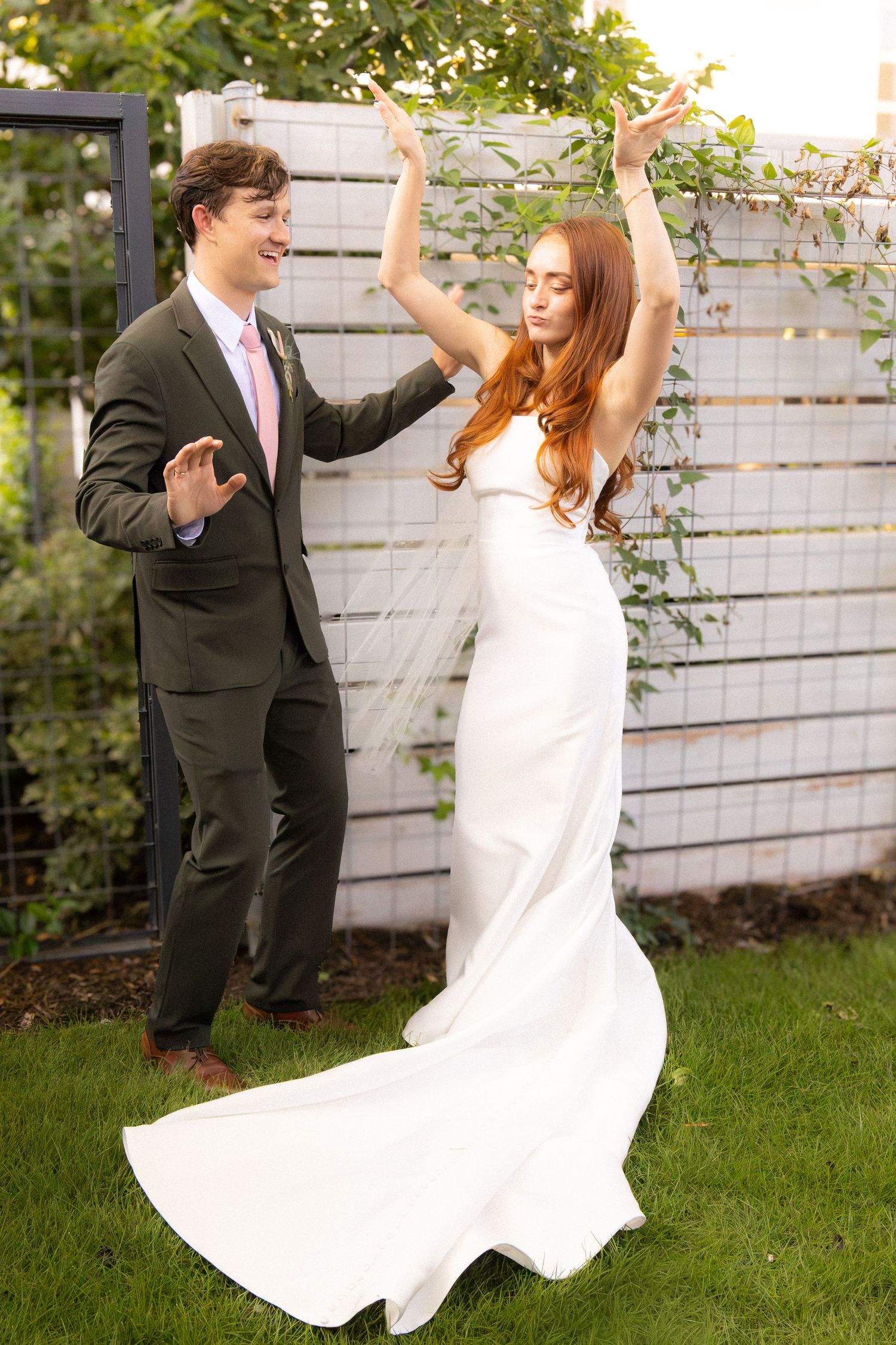 A bride in a white gown dances with a groom in a green suit outdoors on a lush garden lawn.