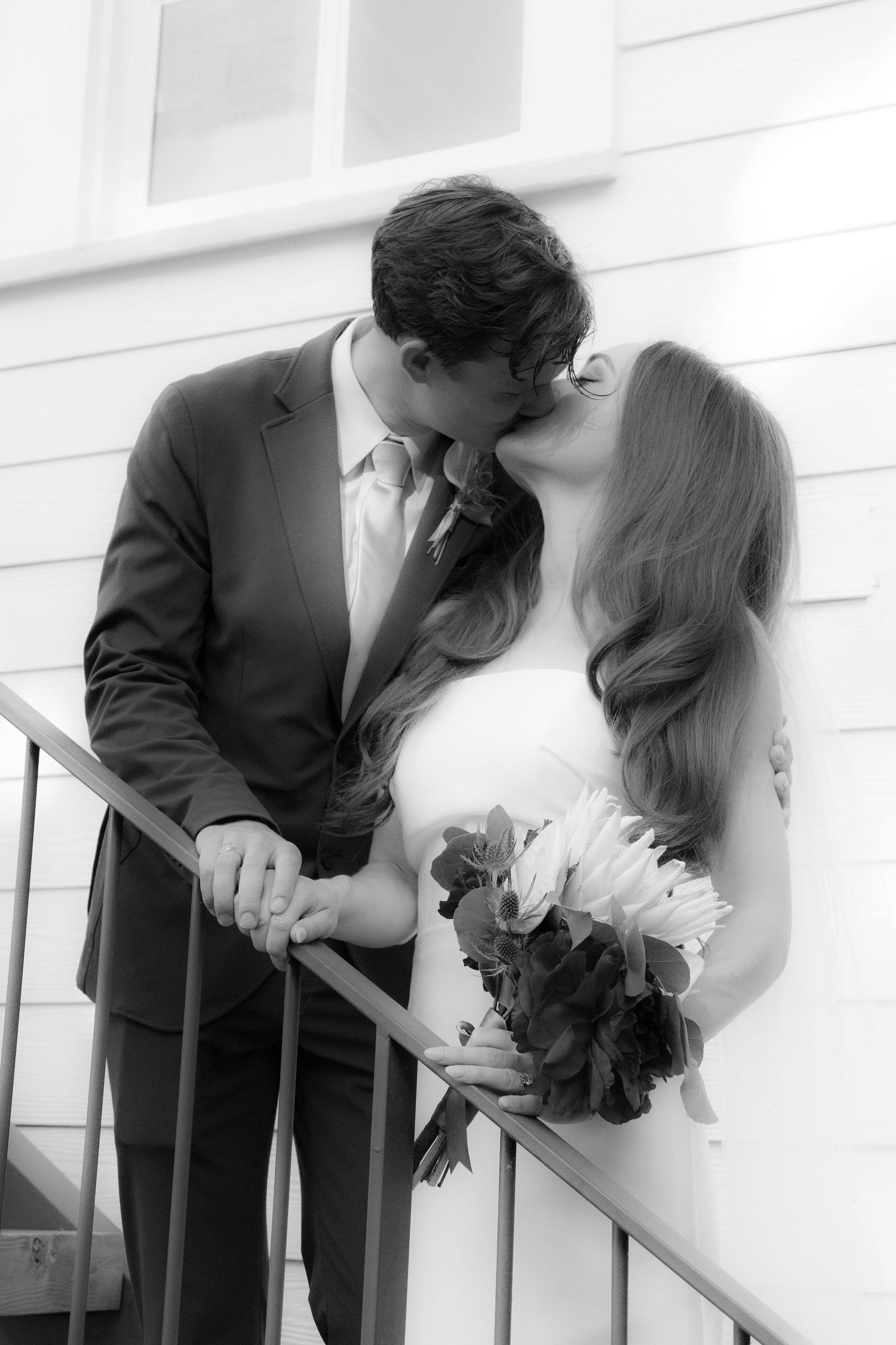 Black and white photo of a bride and groom sharing a kiss on stairs, bride holding a bouquet of flowers.