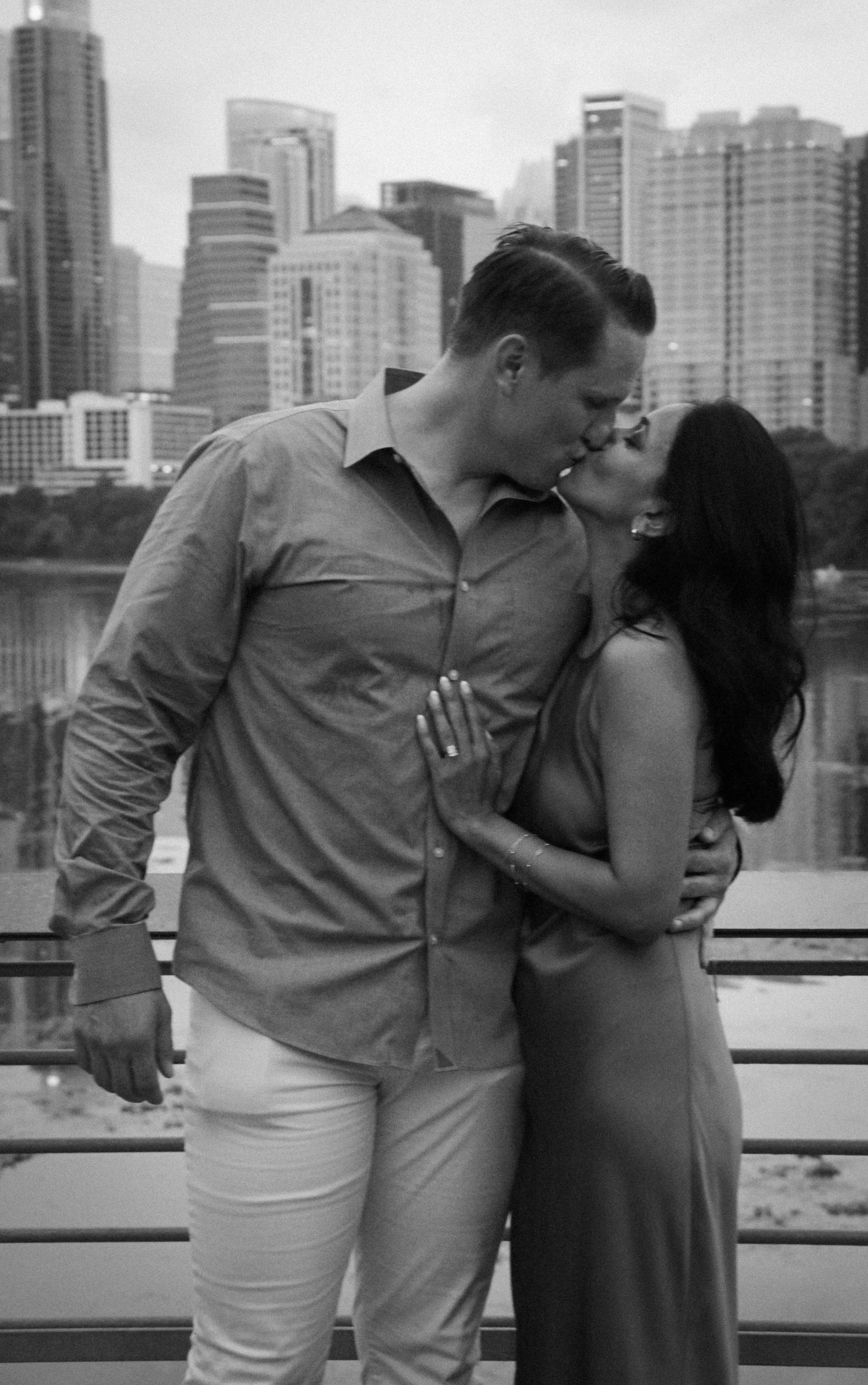Black and white photo of engaged couple sharing a kiss with urban skyline and river in background.
