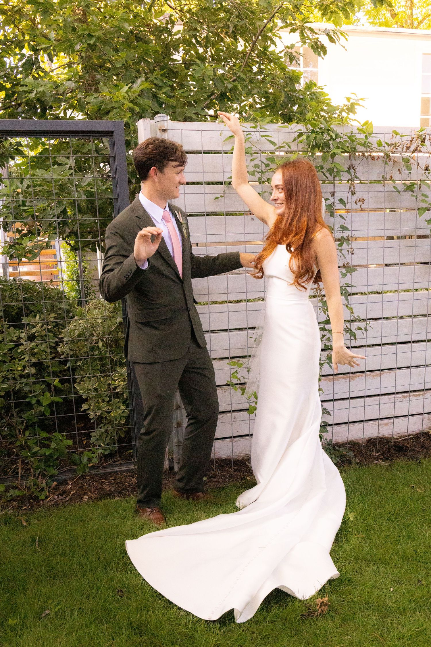A bride in a white gown and groom in a gray suit dance joyfully outdoors on green grass near a garden fence.