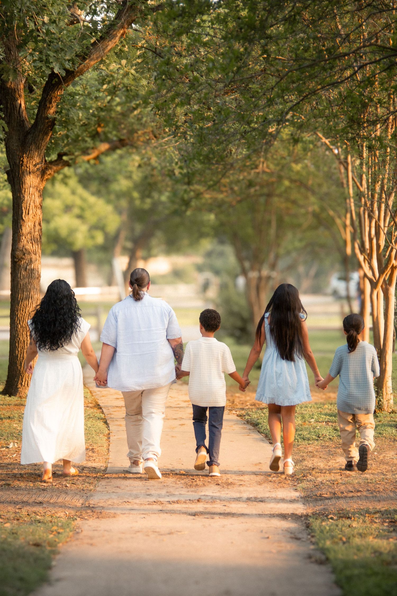 Family of five walking hand-in-hand down a tree-lined park path at golden hour.