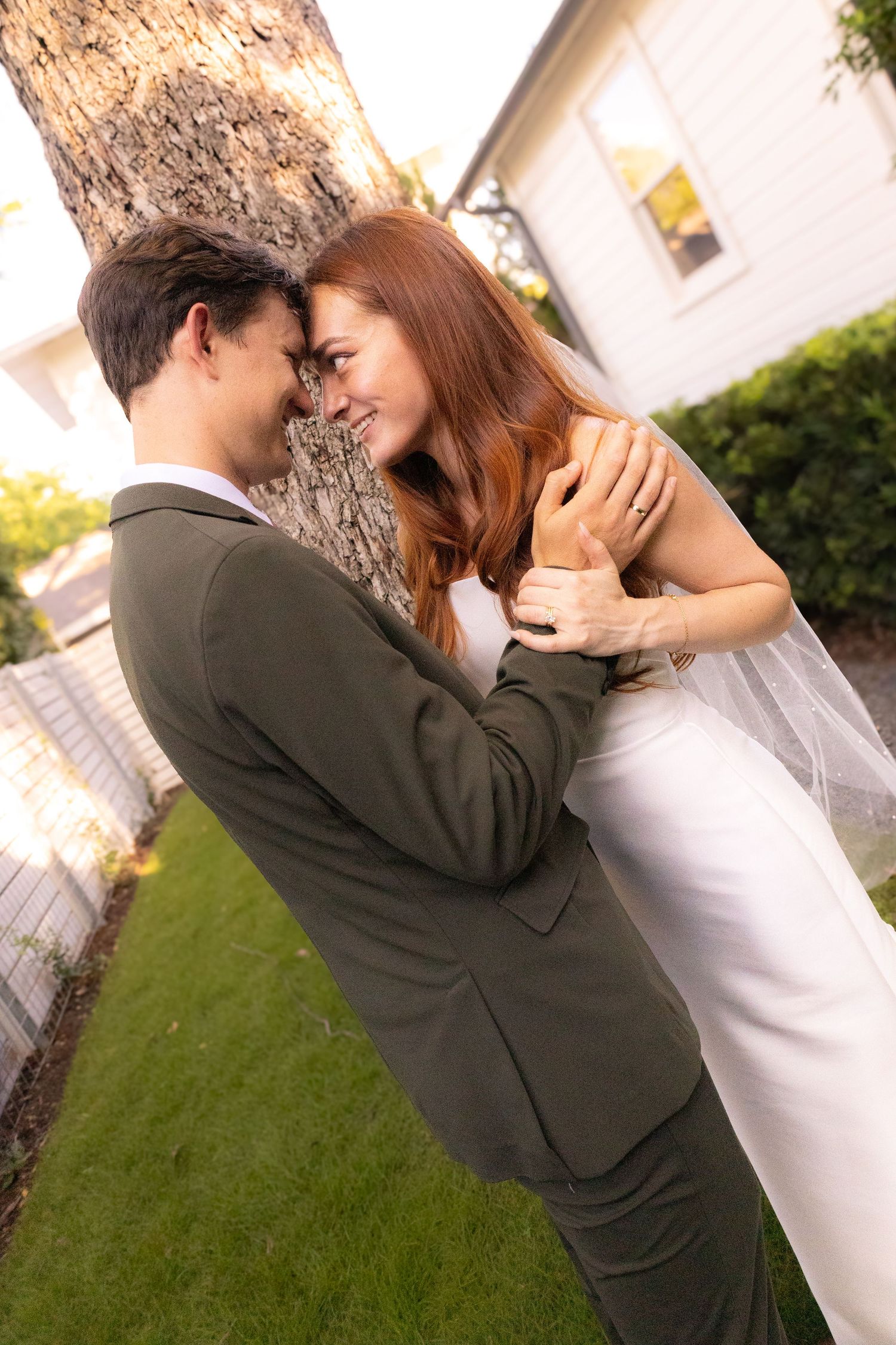 A newlywed couple in wedding attire embraces romantically against a tree in a garden setting.