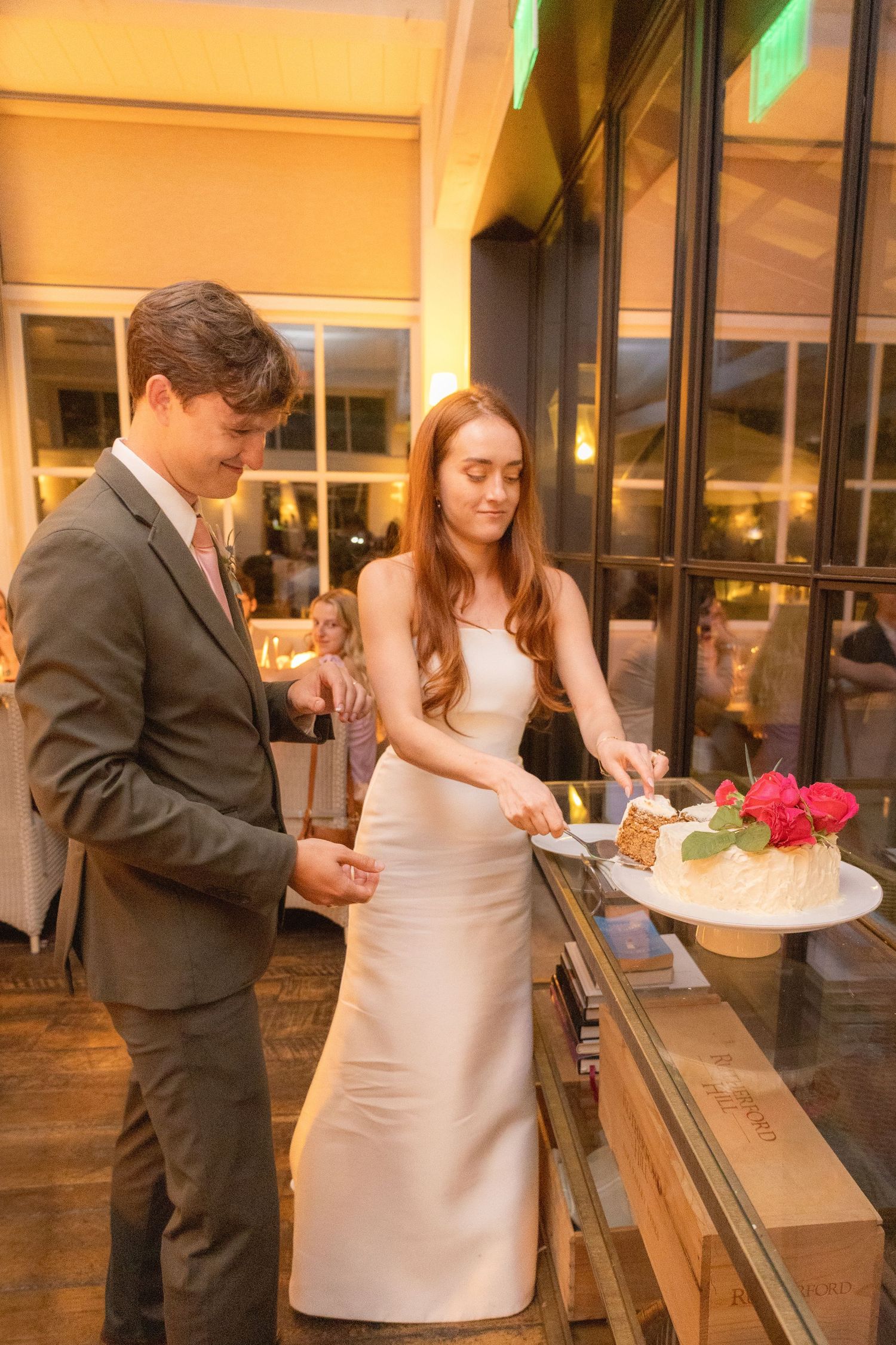 Bride in white gown and groom in gray suit cutting wedding cake together at elegant reception venue.