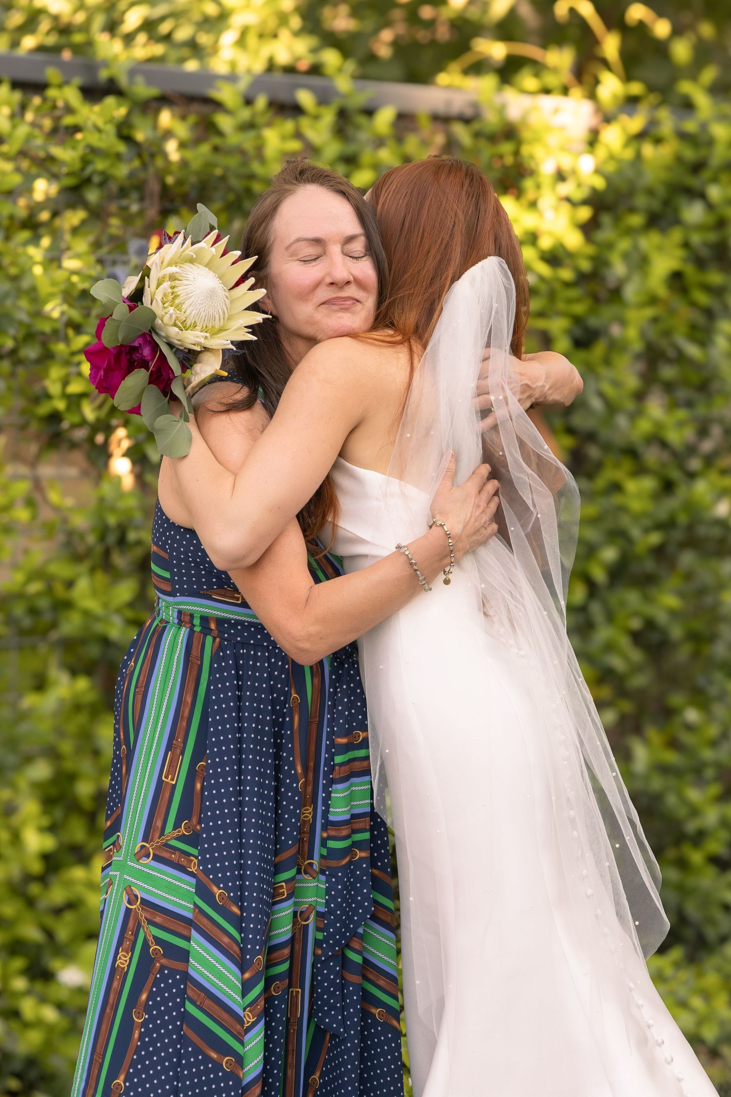 Bride in white gown and veil hugging a woman in a colorful striped dress holding a protea bouquet outdoors.