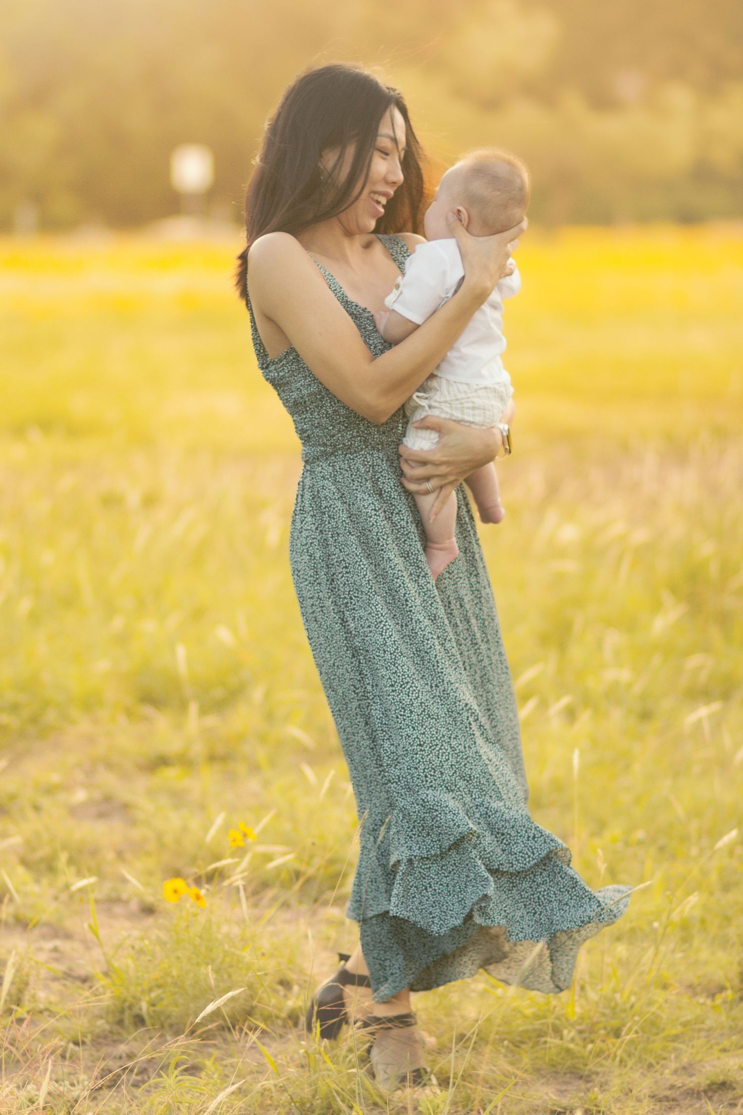 Mother in green floral dress holding infant while standing in golden sunlit meadow.