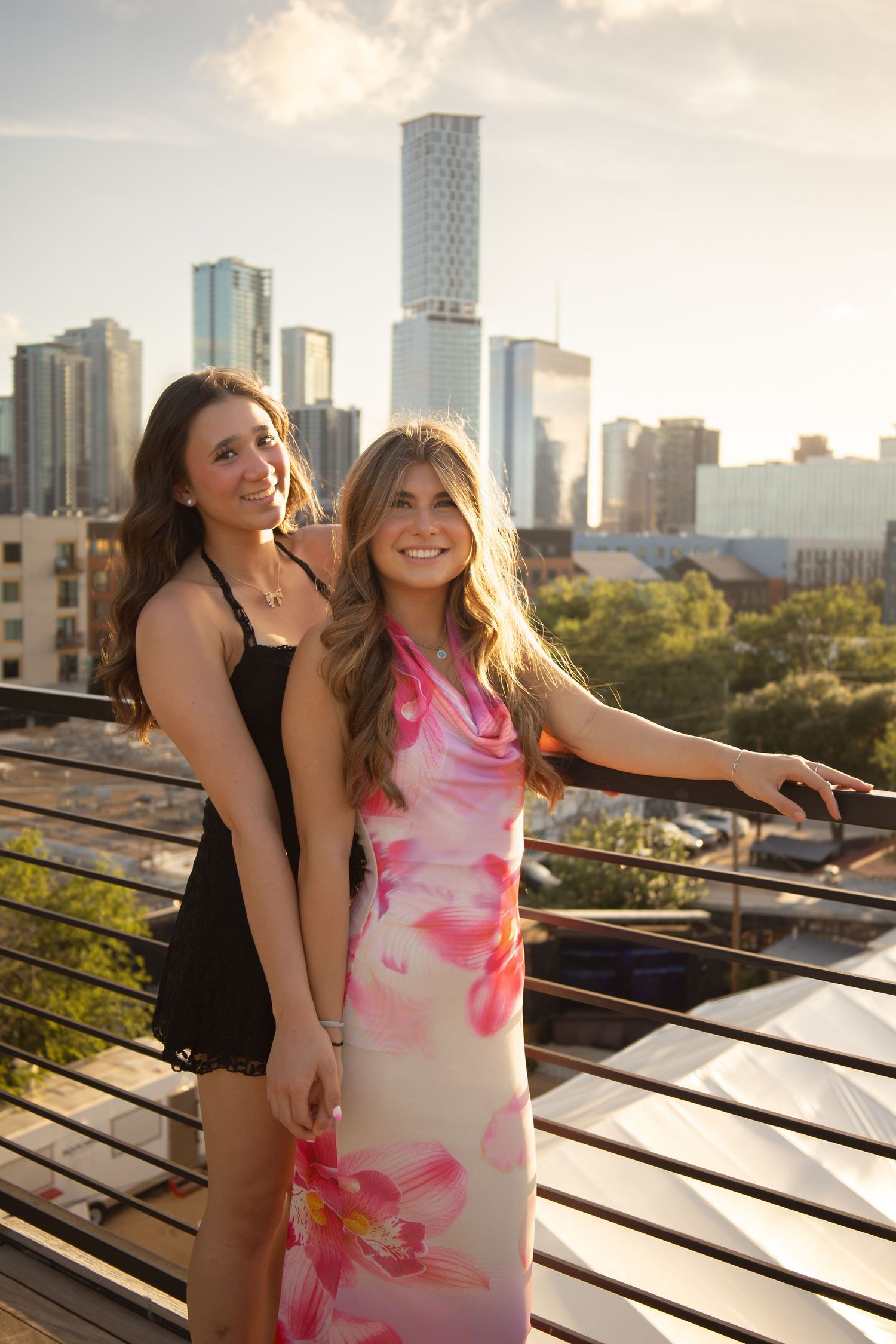 Two smiling women pose on a rooftop terrace with a city skyline backdrop, one in a black dress and one in a floral gown.