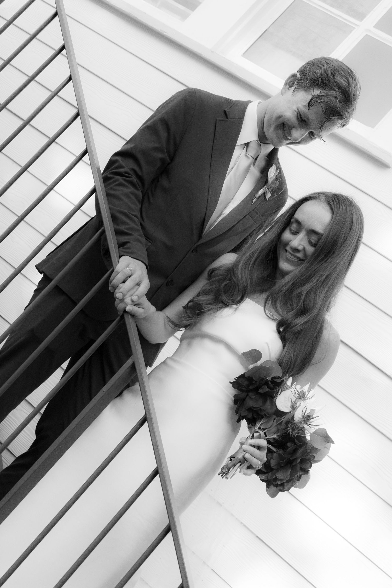 Black and white photo of a bride and groom holding hands on a staircase, bride holding a bouquet.