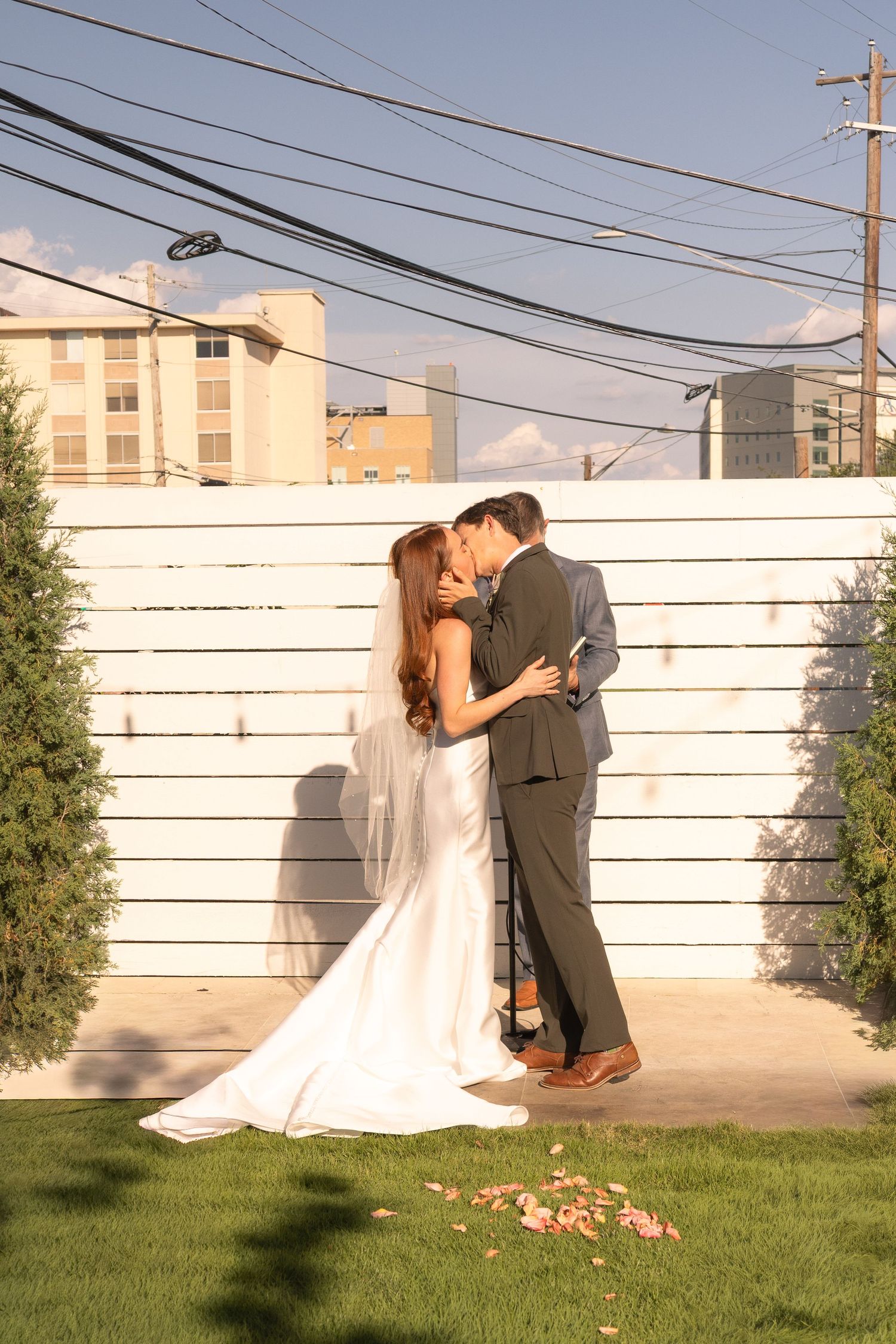 Bride and groom share a romantic kiss in front of a white fence at an outdoor wedding venue.