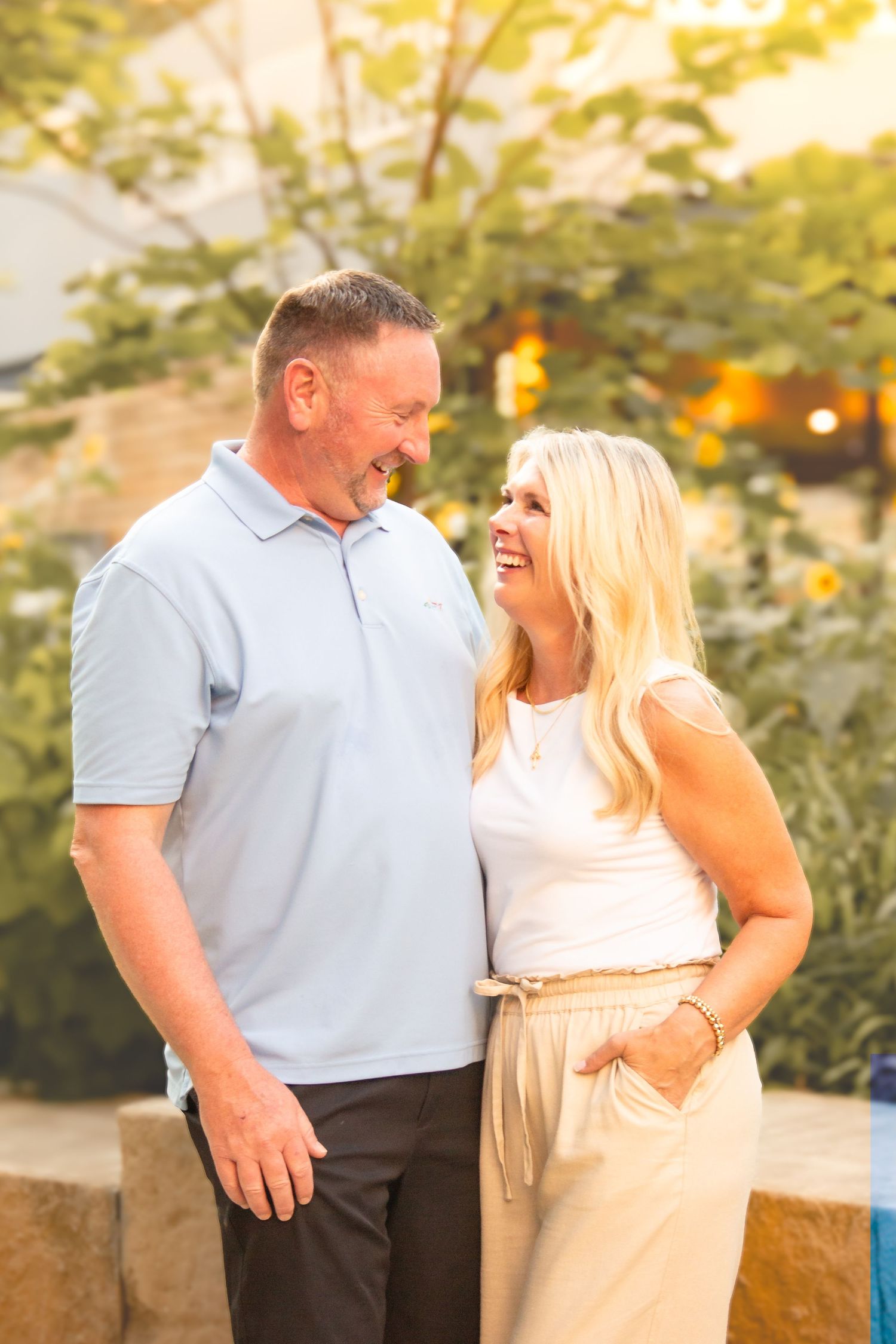 A smiling couple in casual attire share a warm moment outdoors, surrounded by lush greenery and golden sunlight.