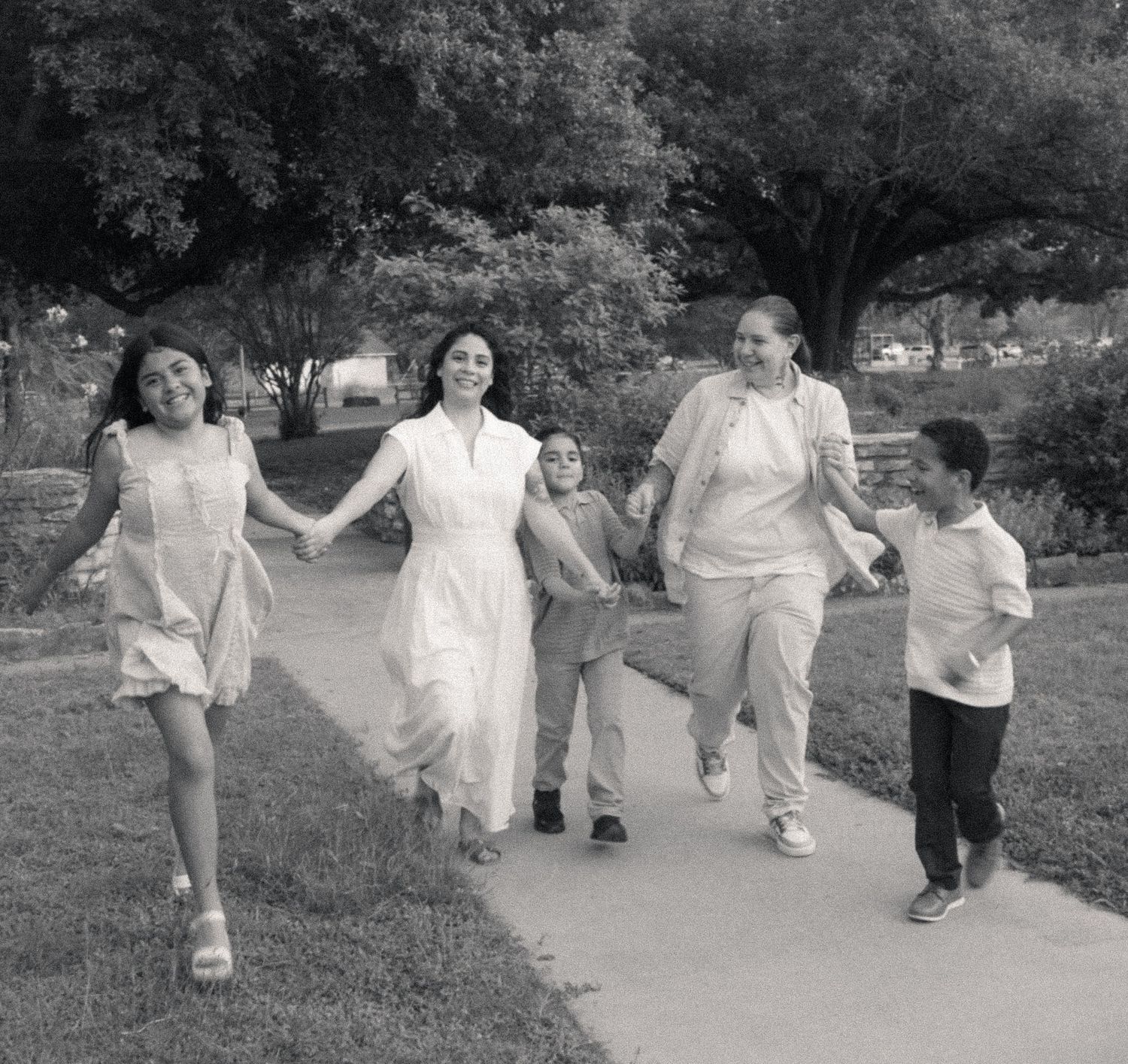 Black and white photo of a joyful family holding hands and walking together on a sunny park pathway.