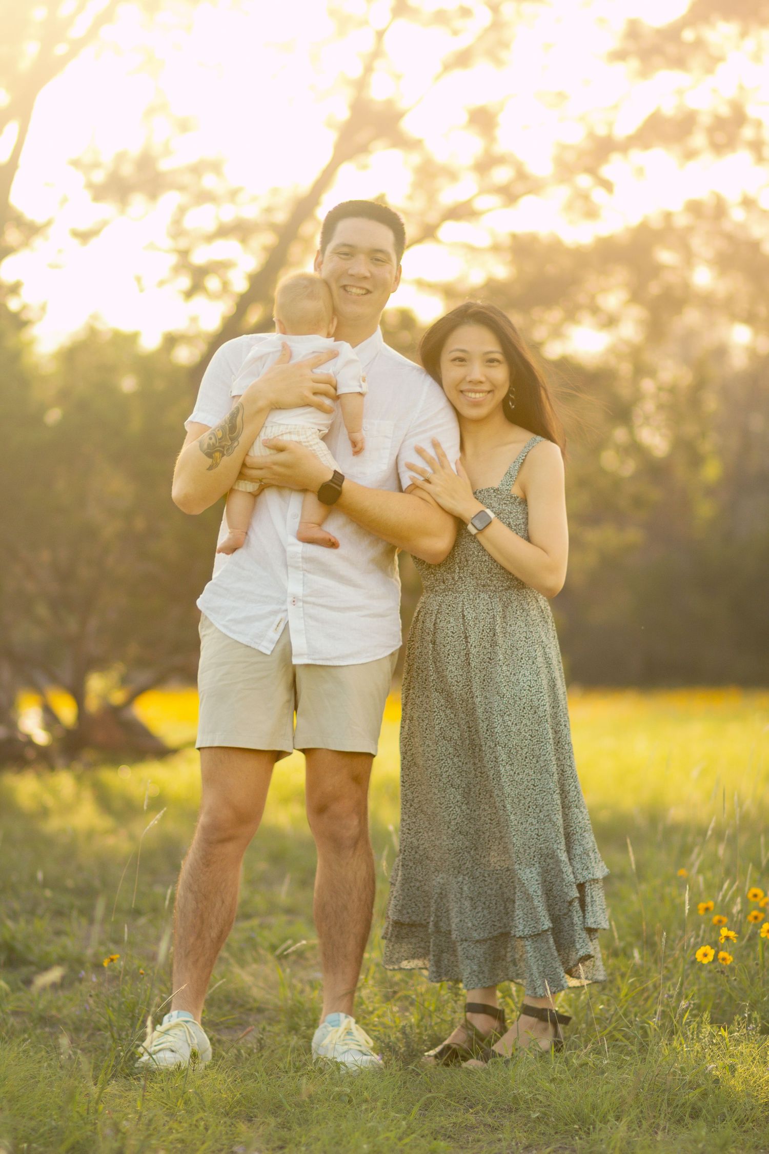A happy young family with a baby poses outdoors in golden hour sunlight surrounded by green grass and trees.