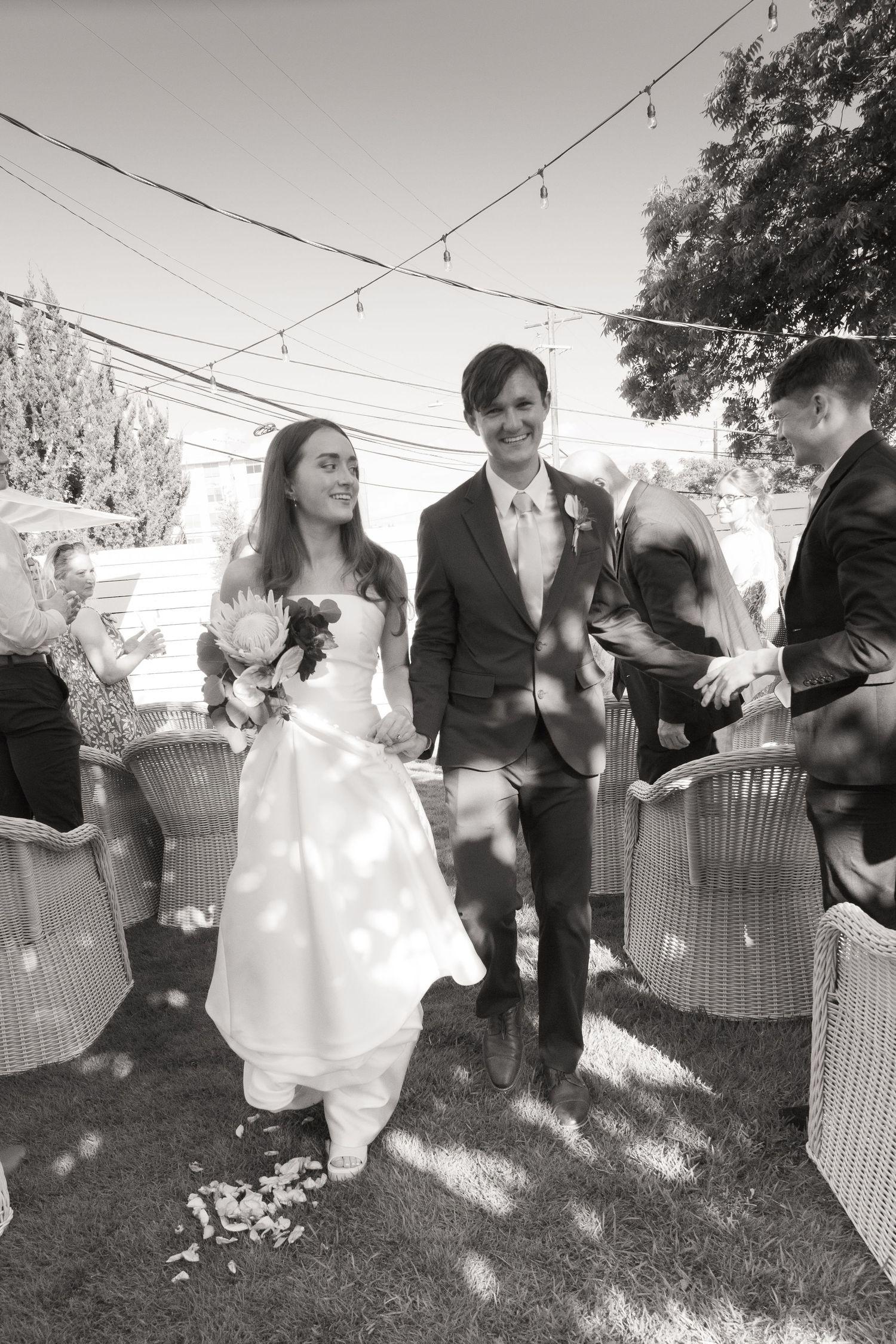 Newlyweds walk down the aisle as guests toss flower petals at an outdoor garden wedding ceremony.