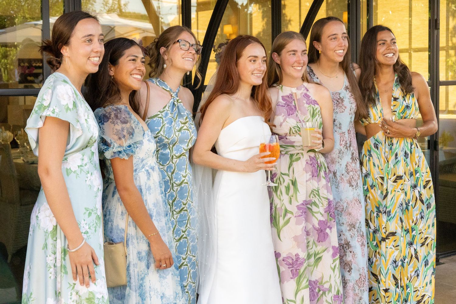 Bride in white strapless gown posing with six bridesmaids in floral dresses outdoors.