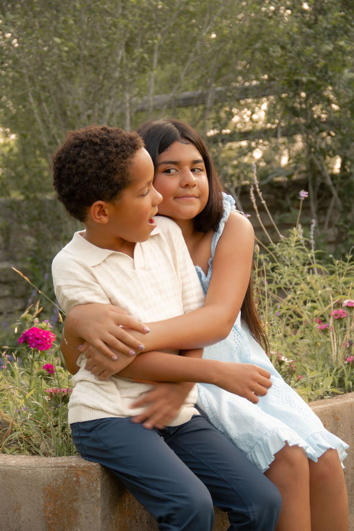 Two children sitting outdoors, a boy and girl hugging affectionately near a garden with pink flowers.