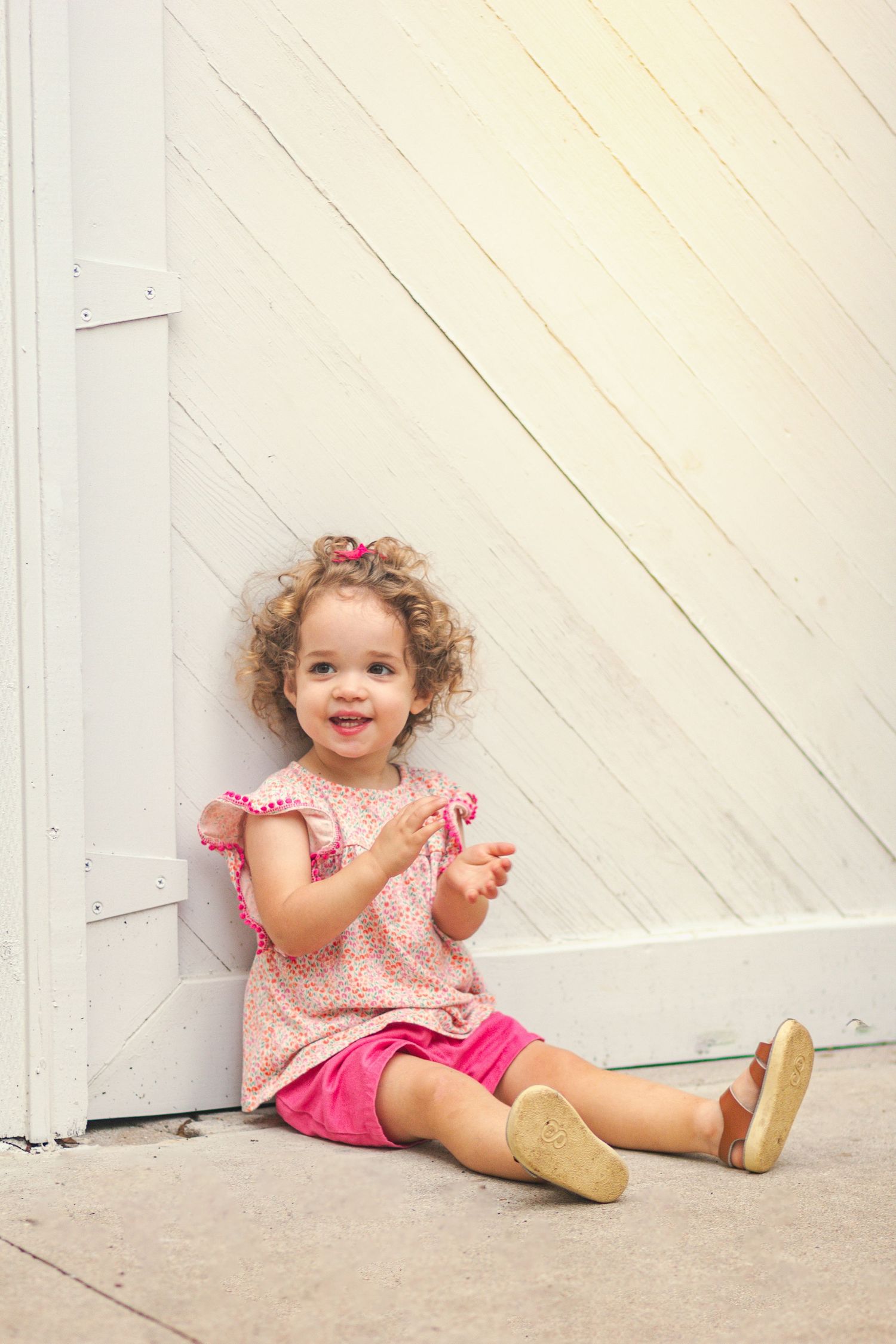 Curly-haired toddler girl in pink dress and shorts sitting on ground, smiling and clapping hands against white wooden wall.