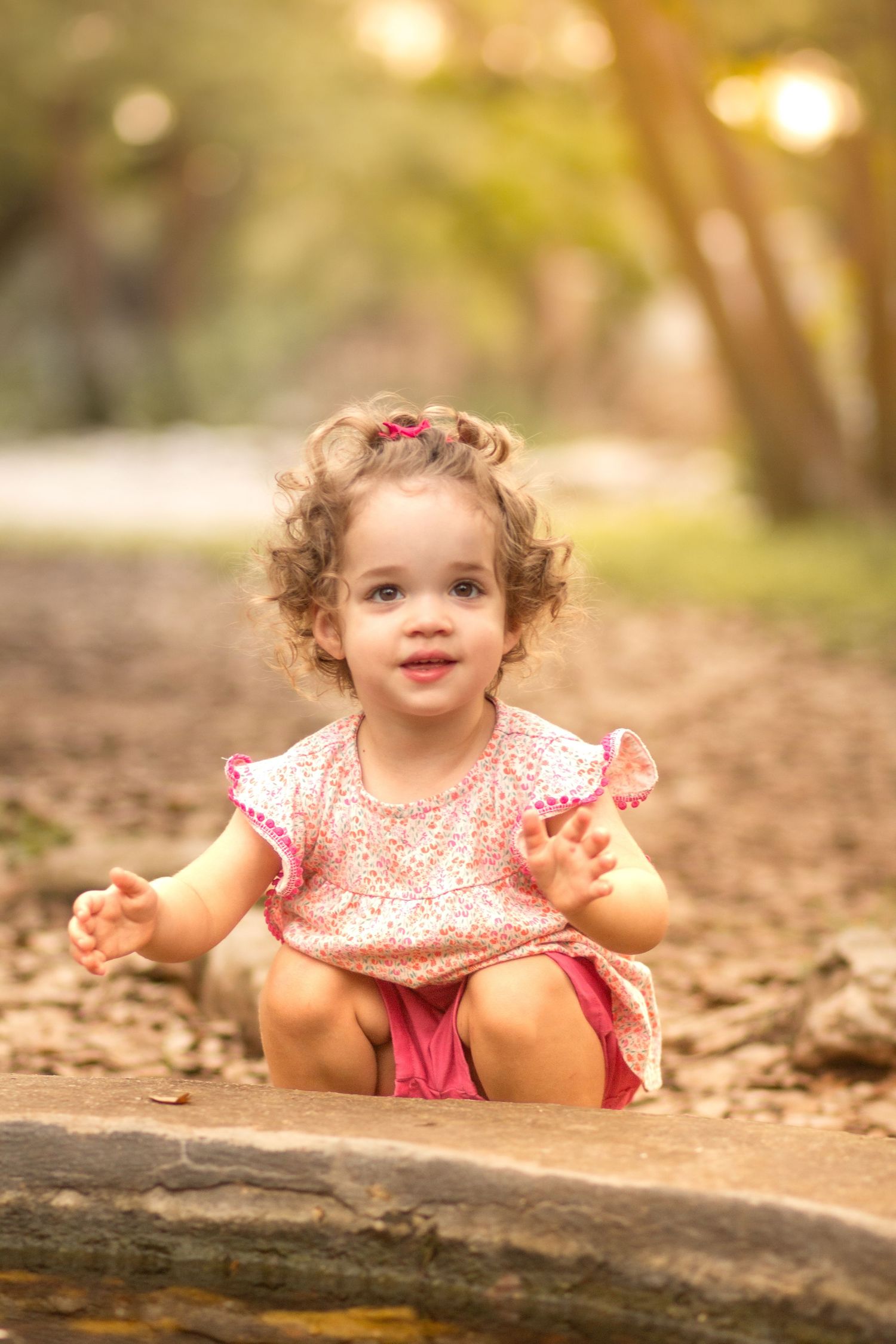 Cute toddler girl in pink floral dress sitting outdoors on a rock, smiling with curly hair in a warm park setting.