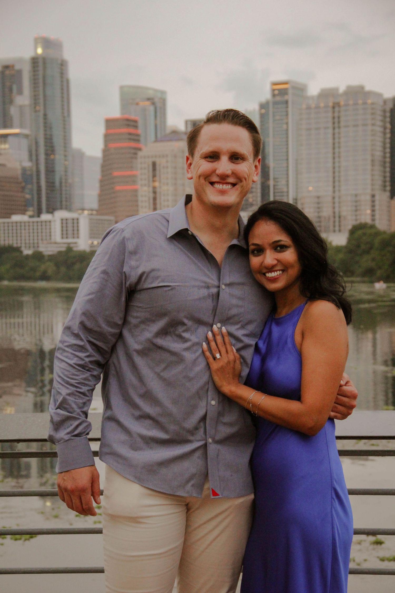 Happy engaged couple posing together with ring visible against city skyline backdrop.