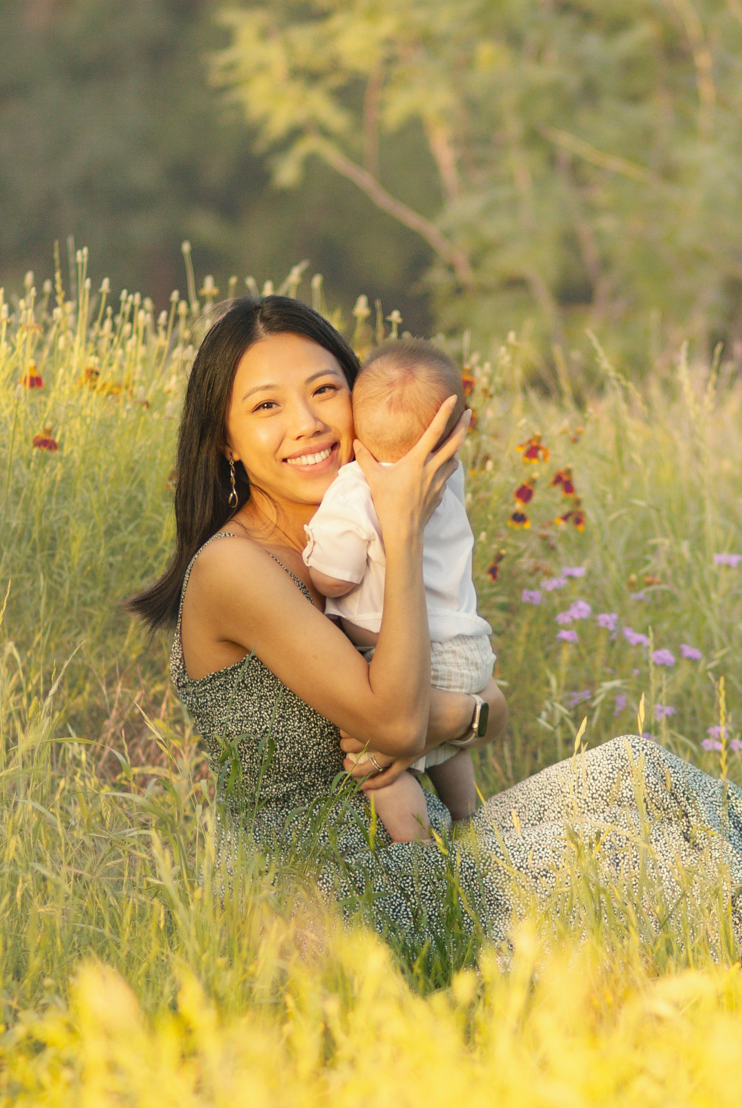 Smiling Asian mother holding baby in sunny wildflower meadow, wearing floral dress, golden hour outdoor family portrait.